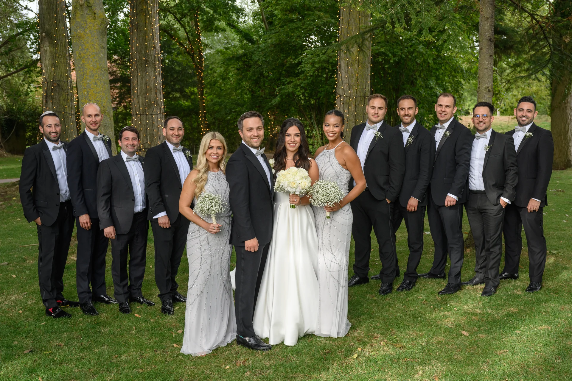 The bride and groom pose happily alongside their large wedding party of groomsmen in black tuxedos and bridesmaids in grey beaded gowns outdoors near trees strung with fairy lights.