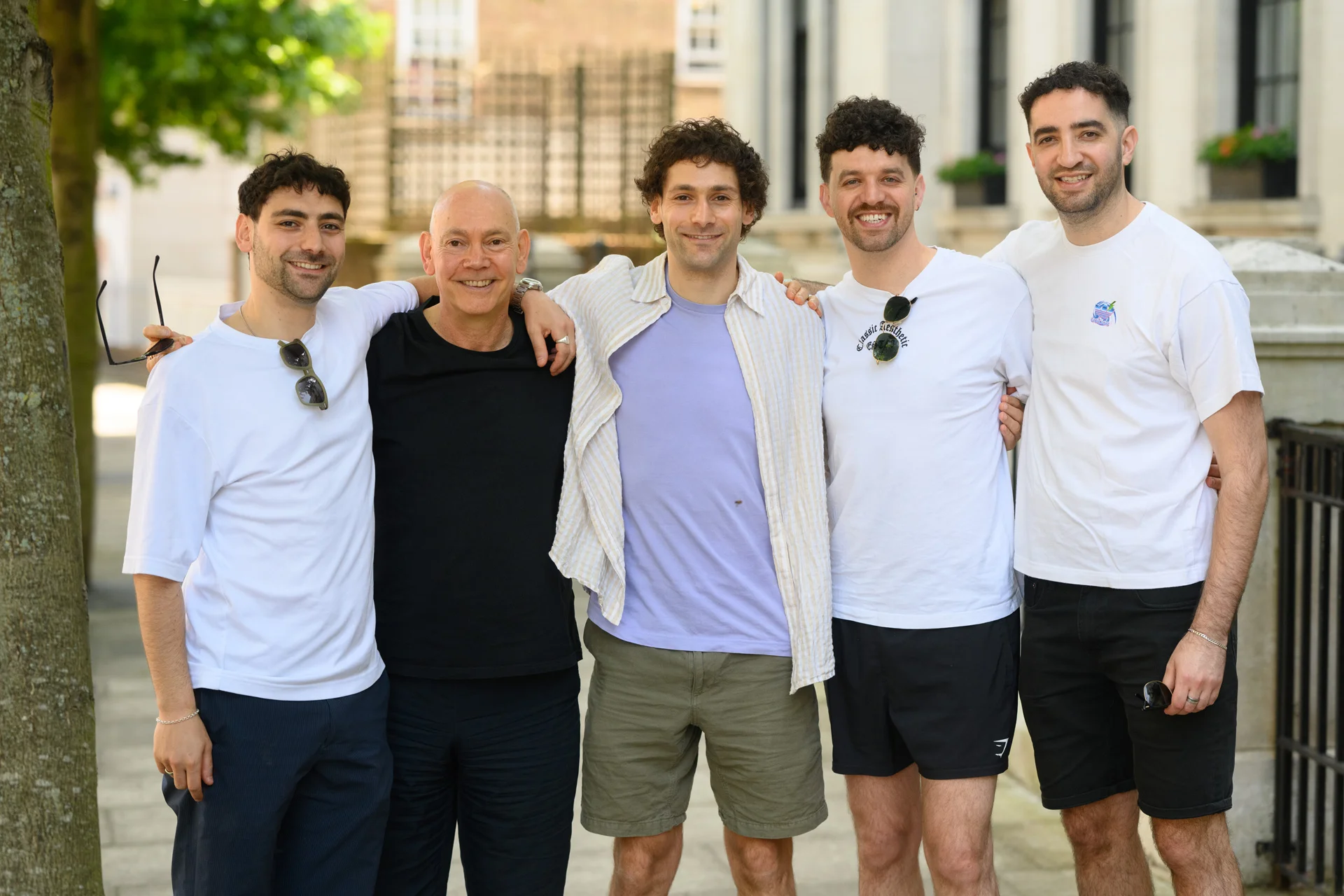 A group of five men stand outdoors in a city setting, smiling and posing with their arms around each other's shoulders.