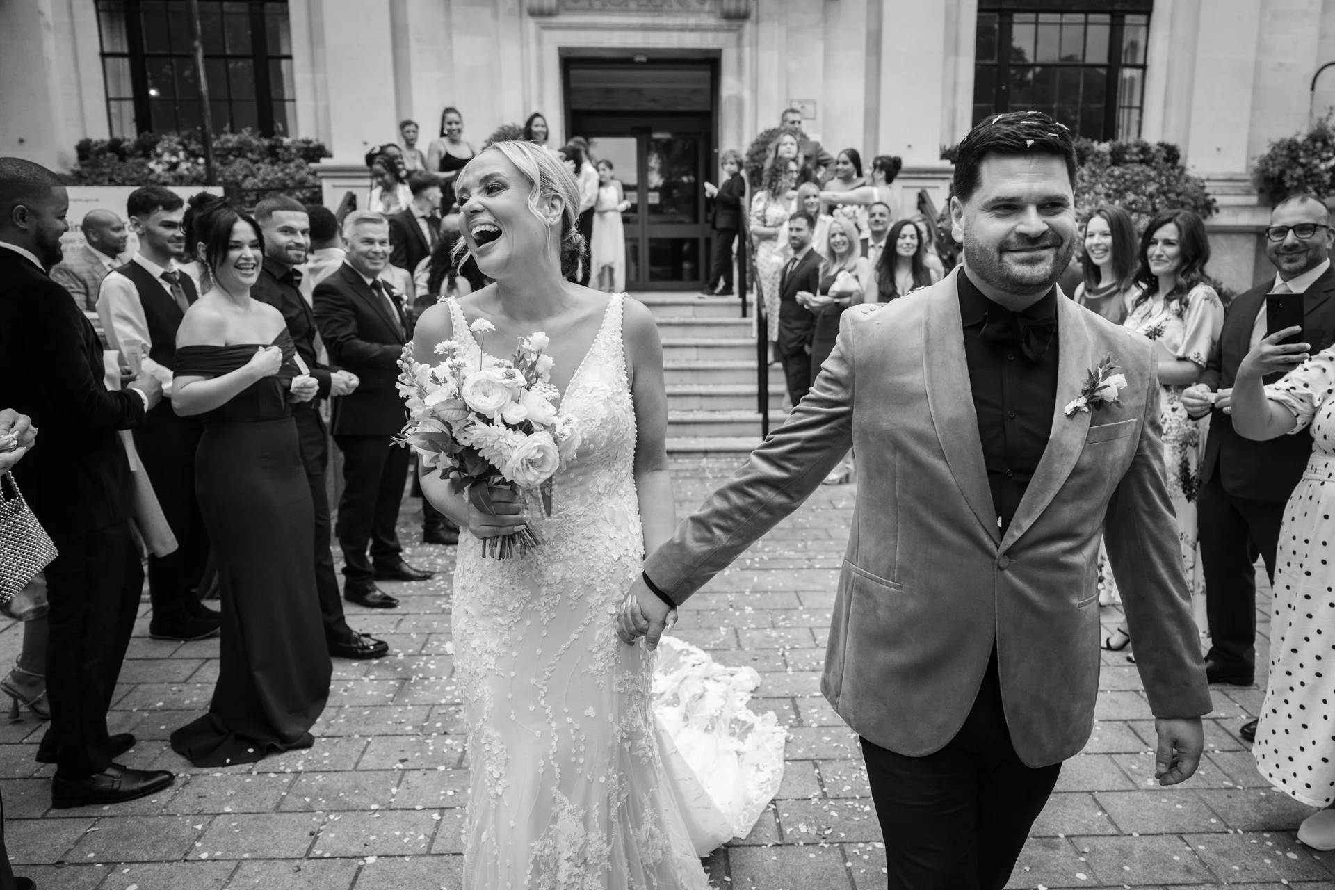 Newlyweds walk hand in hand down the stone steps of a town hall while laughing joyfully as guests cheer and shower them with confetti.