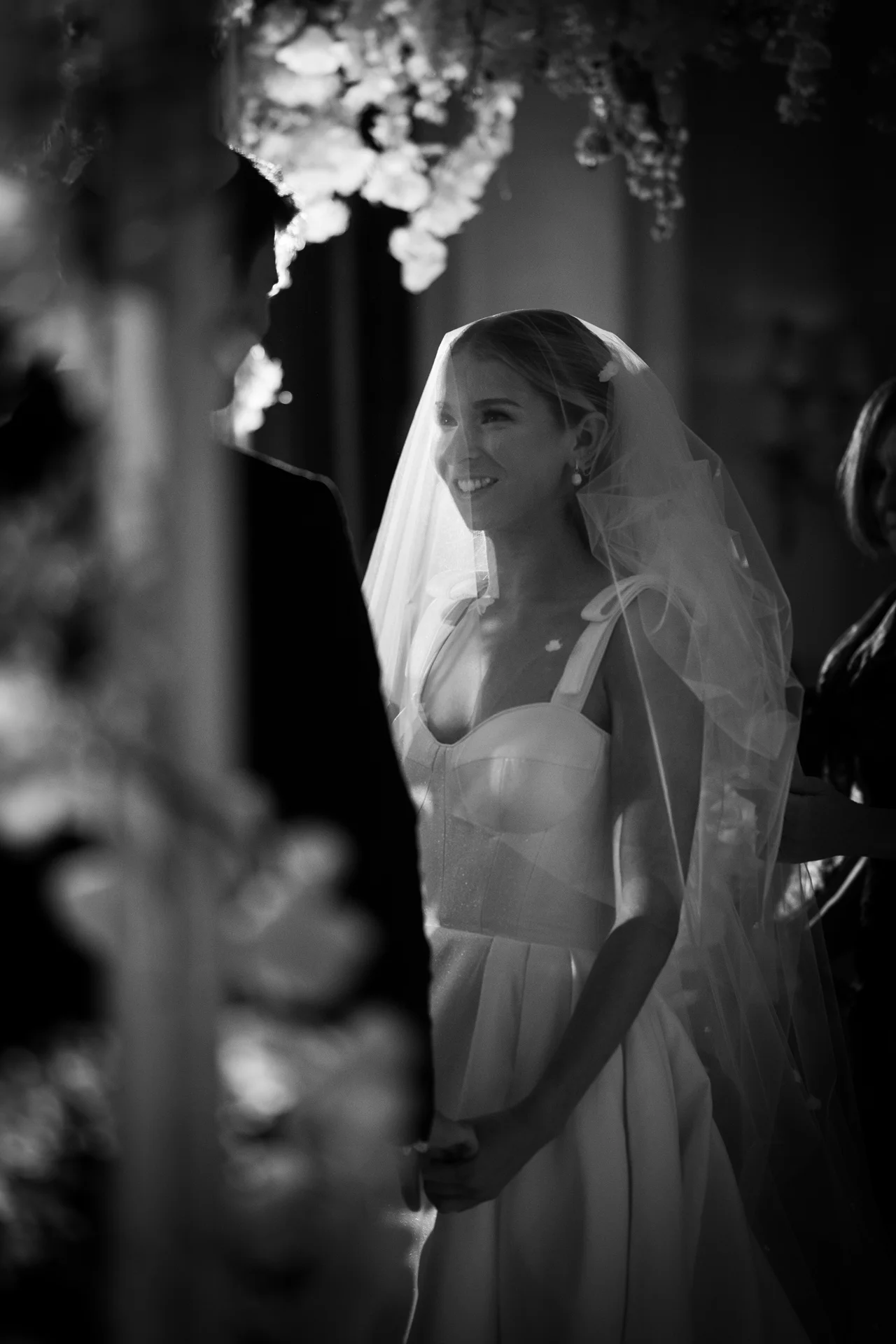 A smiling bride wearing a veil and structured gown looks lovingly toward the groom during their wedding ceremony.