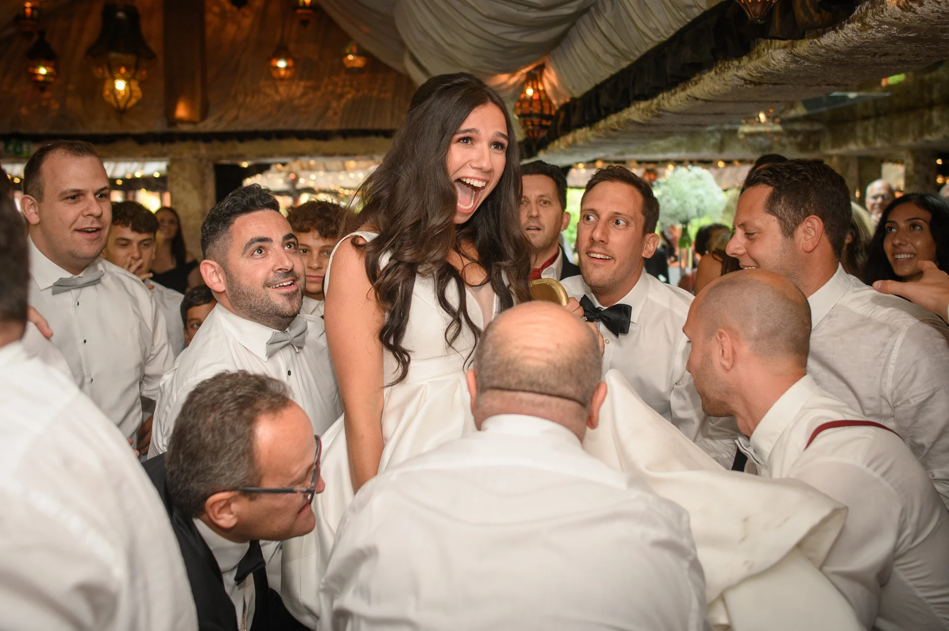 A joyful bride is lifted high into the air by wedding guests during an energetic reception celebration.