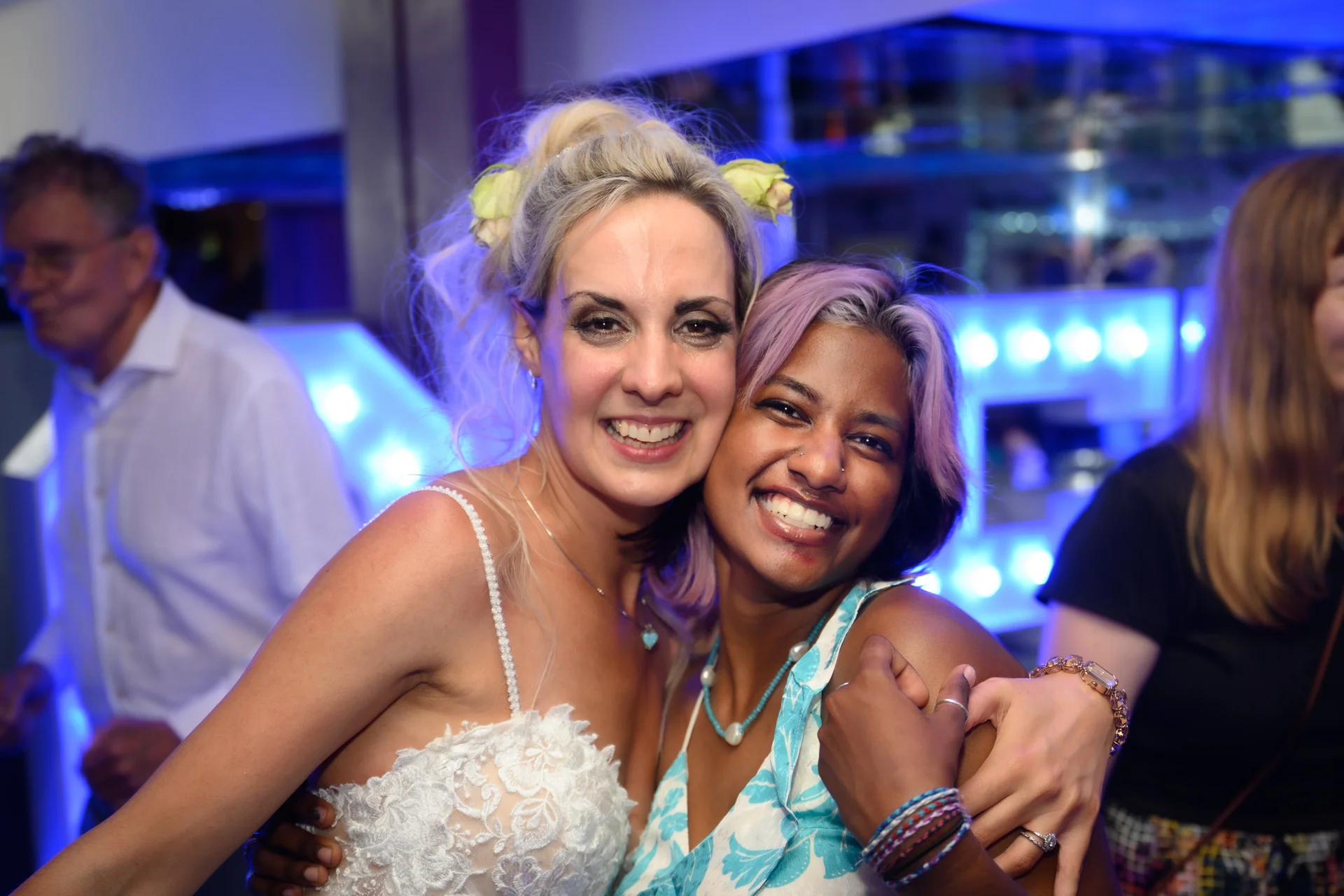 Two smiling brides embrace joyfully at an evening wedding reception illuminated by vibrant blue party lights.
