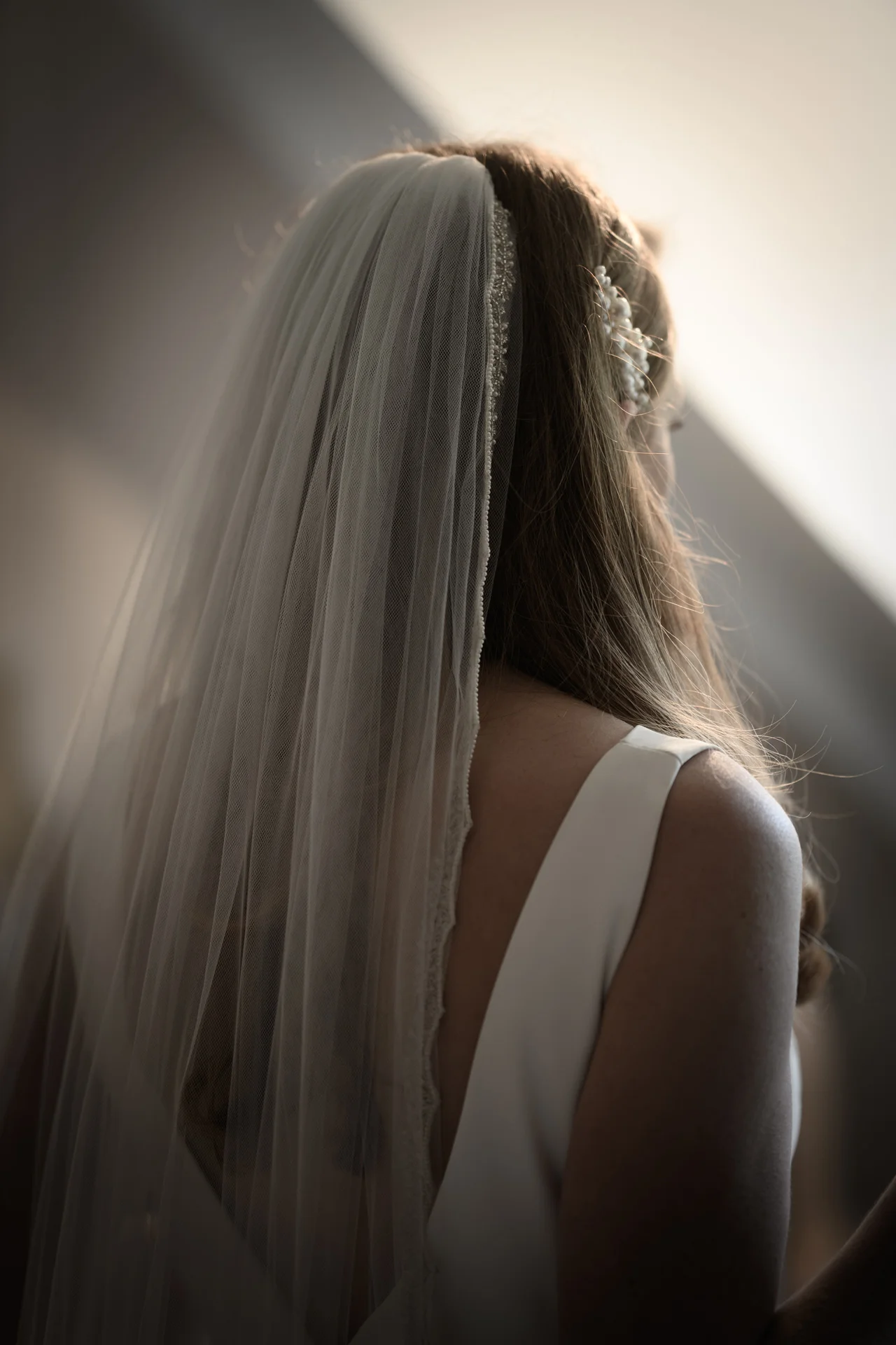 A close-up back view of a bride wearing a simple white crepe gown, a beaded chapel veil, and a delicate pearl hair comb illuminated by soft window light.
