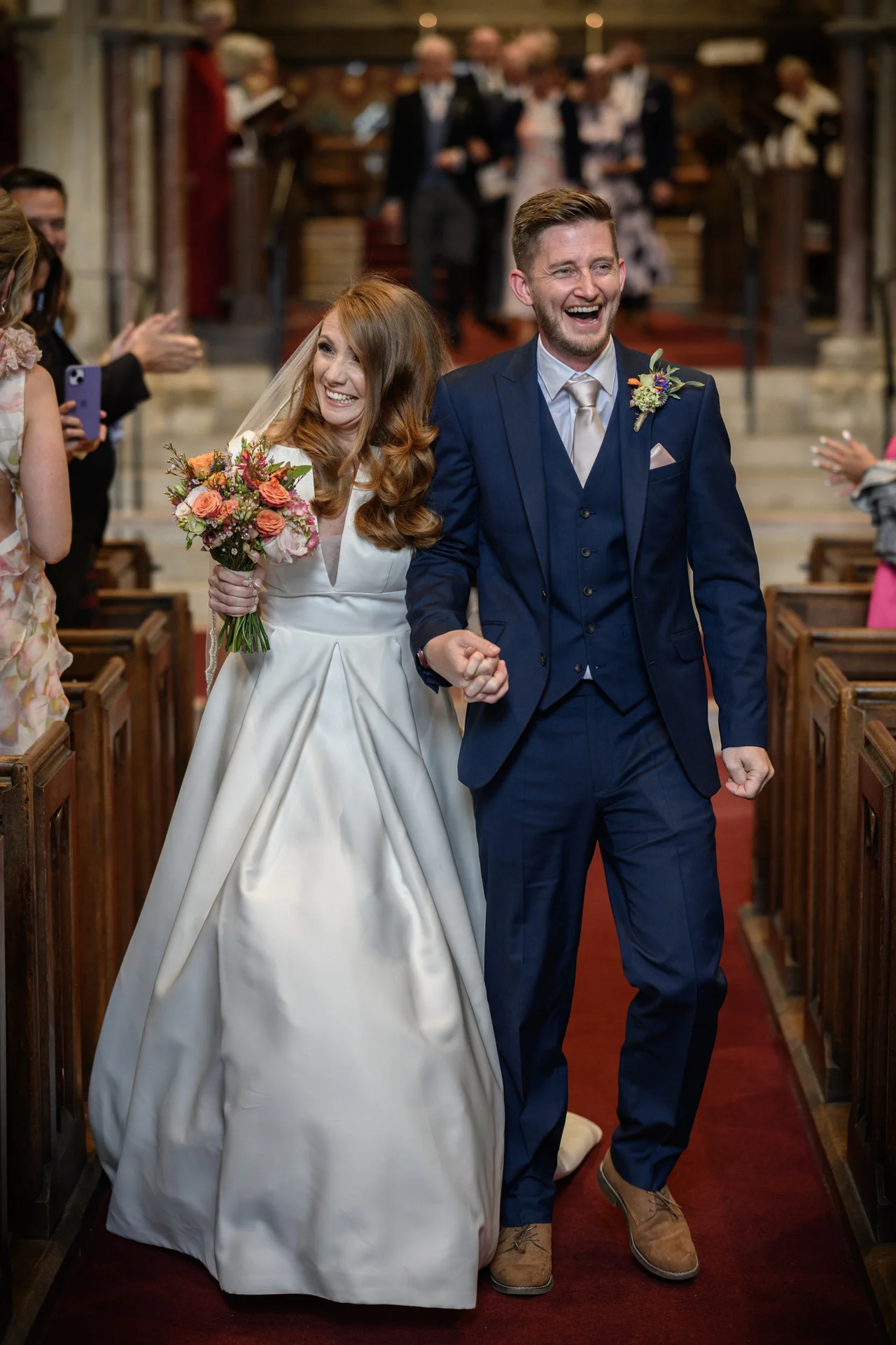 A joy-filled newlywed couple laughs happily as they walk arm in arm down the red-carpeted aisle of a traditional church following their ceremony.