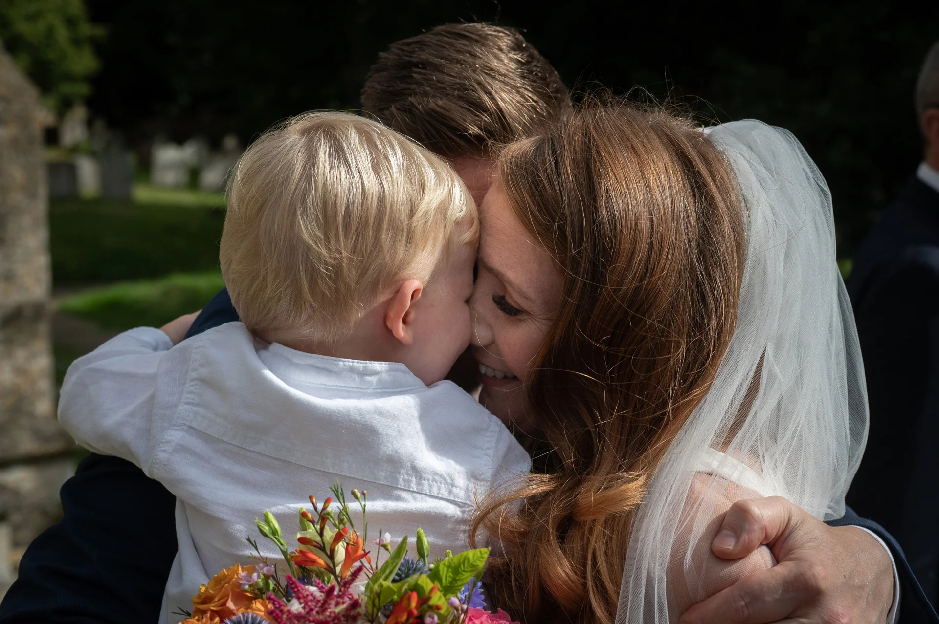 A bride and groom share a tender, close-up embrace with a young blonde child while holding a colourful wedding bouquet outdoors.