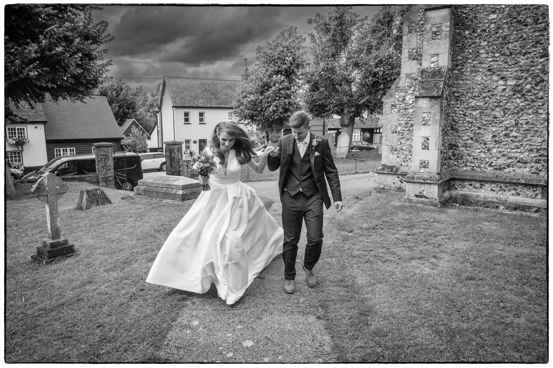 A newlywed couple run hand in hand down the path outside an old stone church under a dramatic, cloudy sky.