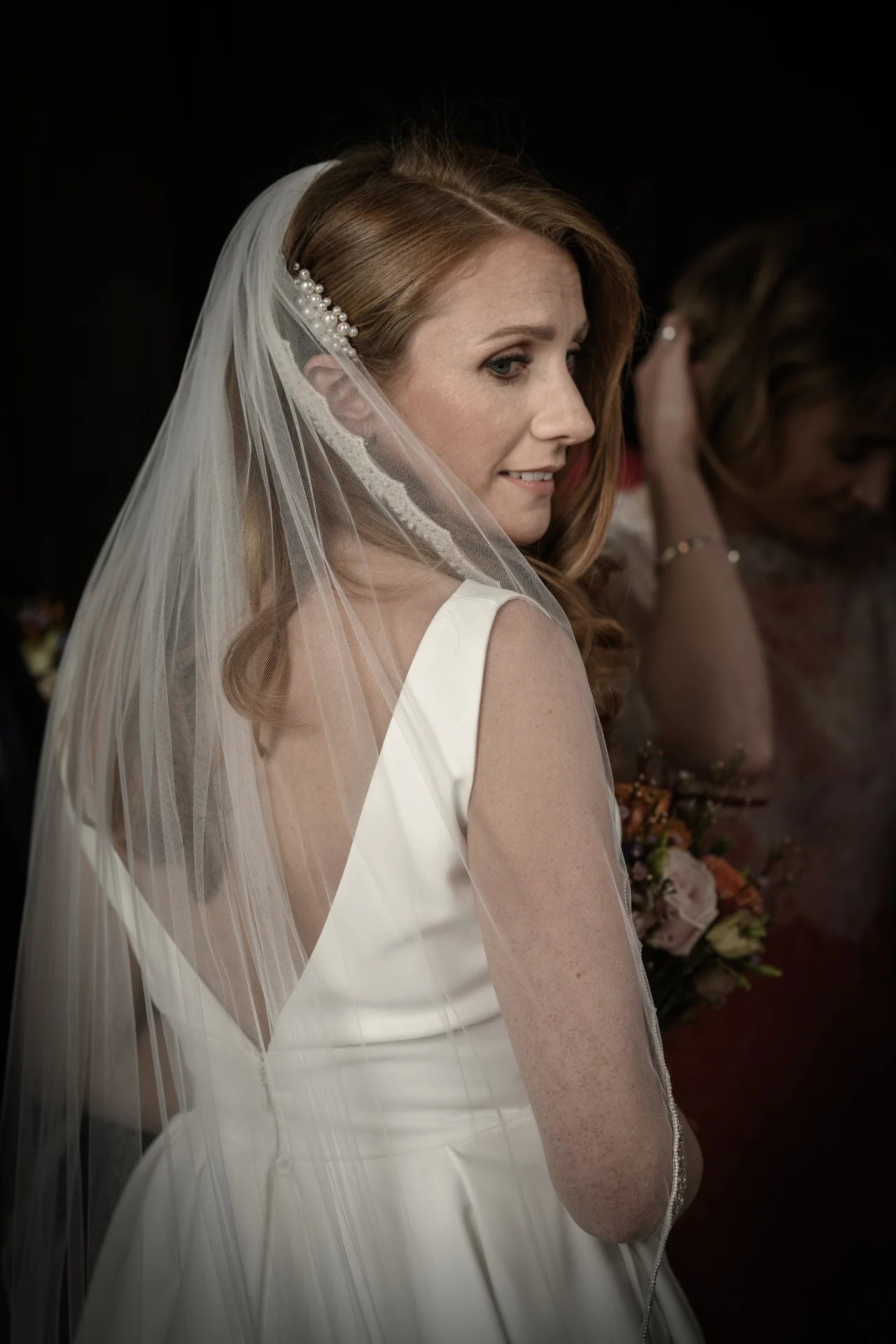 A stunning red-haired bride wearing a satin v-neck gown and a delicate veil secured with a pearl hair comb looks back over her shoulder.