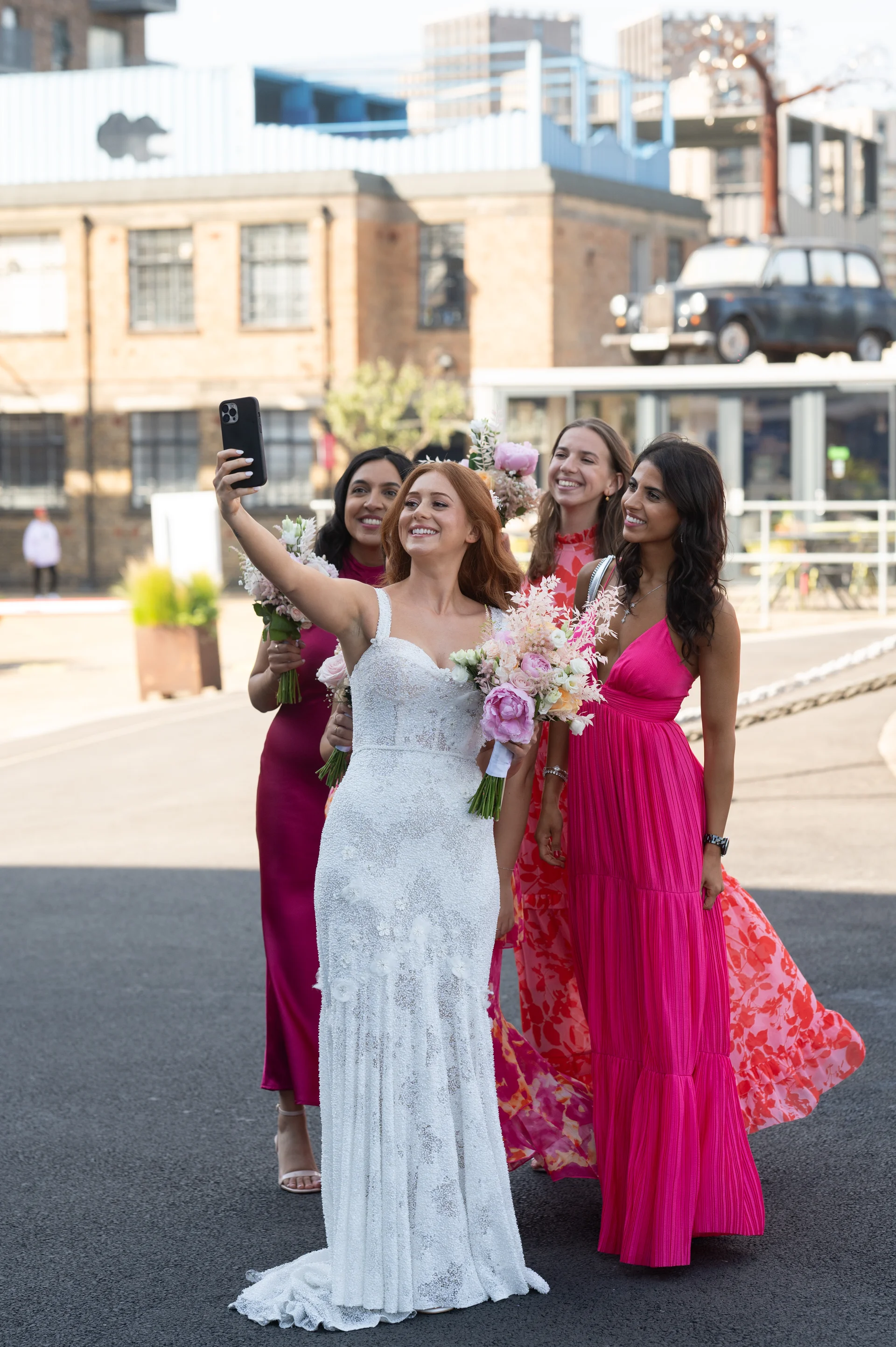 A radiant red-haired bride wearing a beaded white wedding dress holds a bouquet and takes a selfie with her smiling bridesmaids in vibrant pink dresses outside an urban venue.