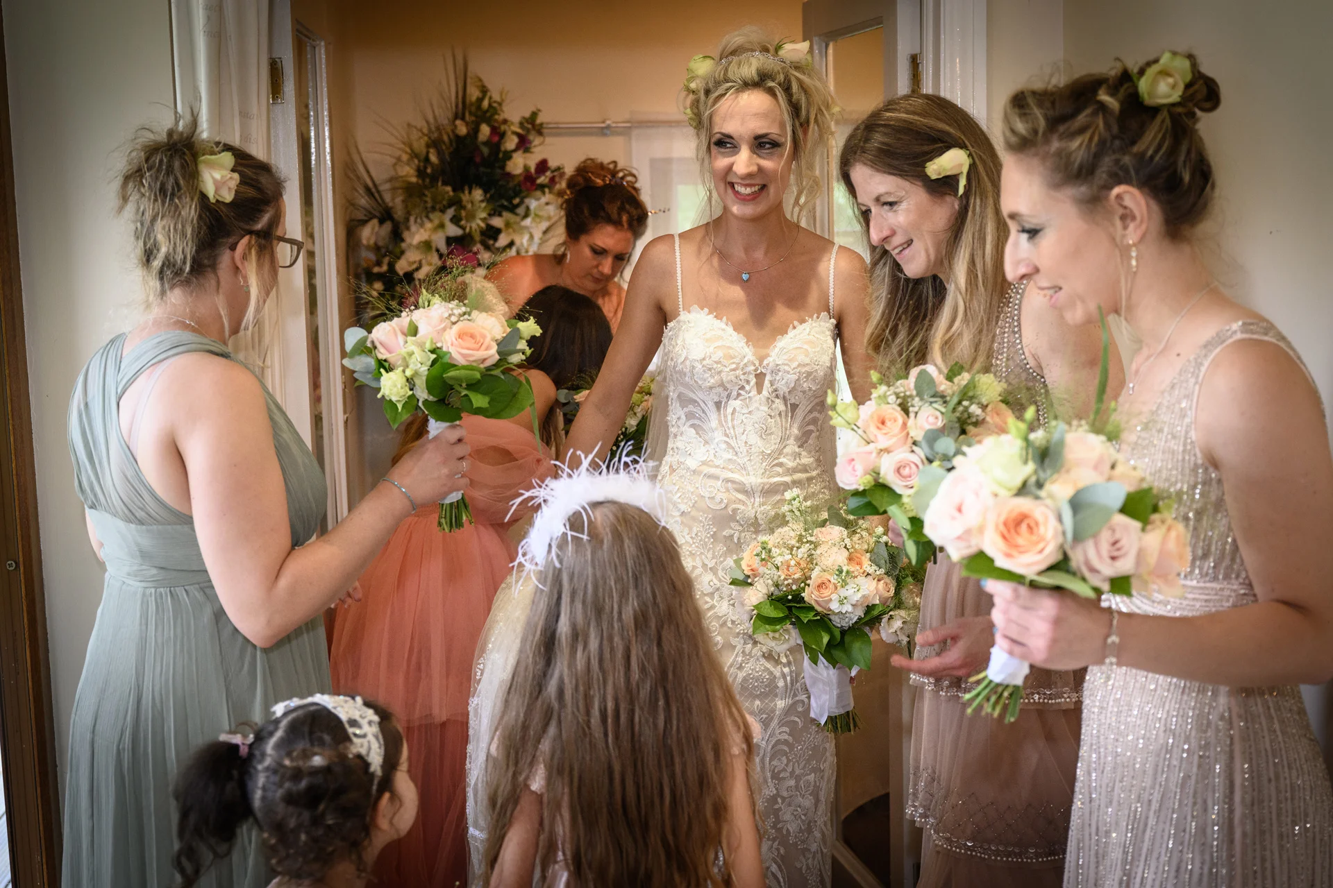 A smiling bride holding a floral bouquet shares a joyful first look with her bridesmaids and flower girls during morning bridal preparations.