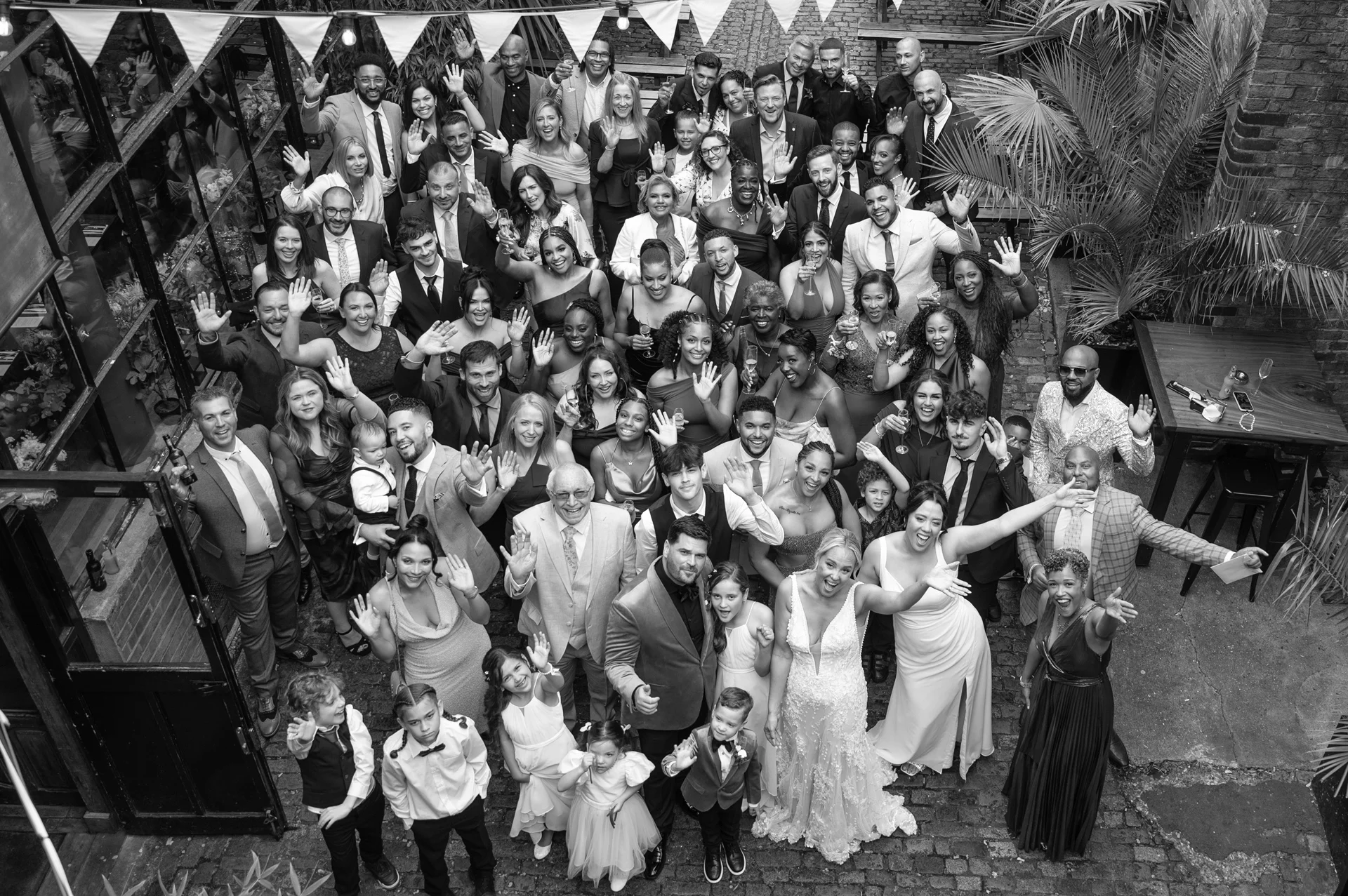An overhead drone shot captures a large wedding crowd waving and celebrating together in a brick courtyard adorned with bunting.
