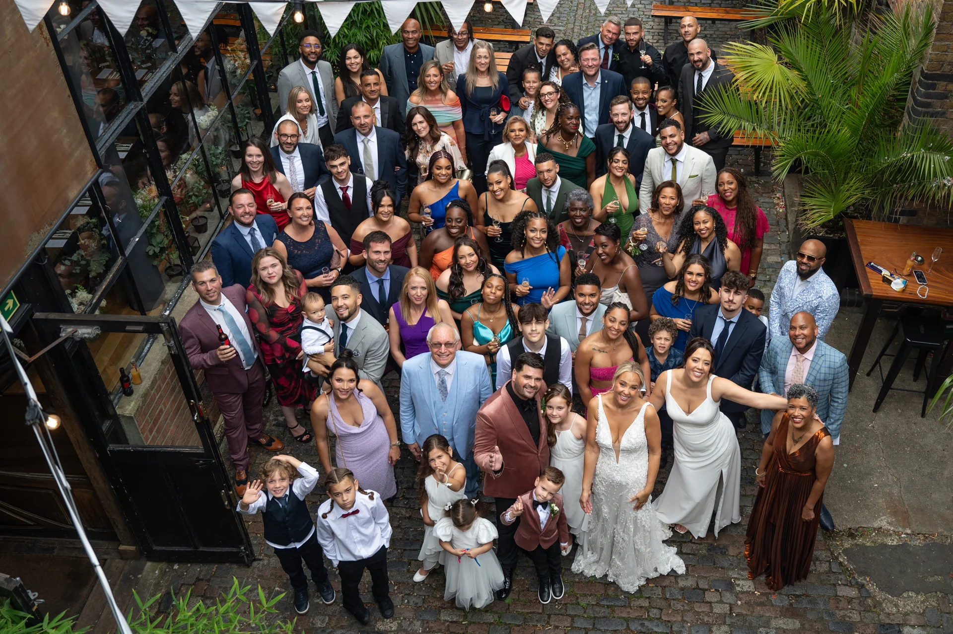 An overhead high-angle group photograph capturing a large wedding party and guests gathered in a cobbled courtyard during the daytime celebration.