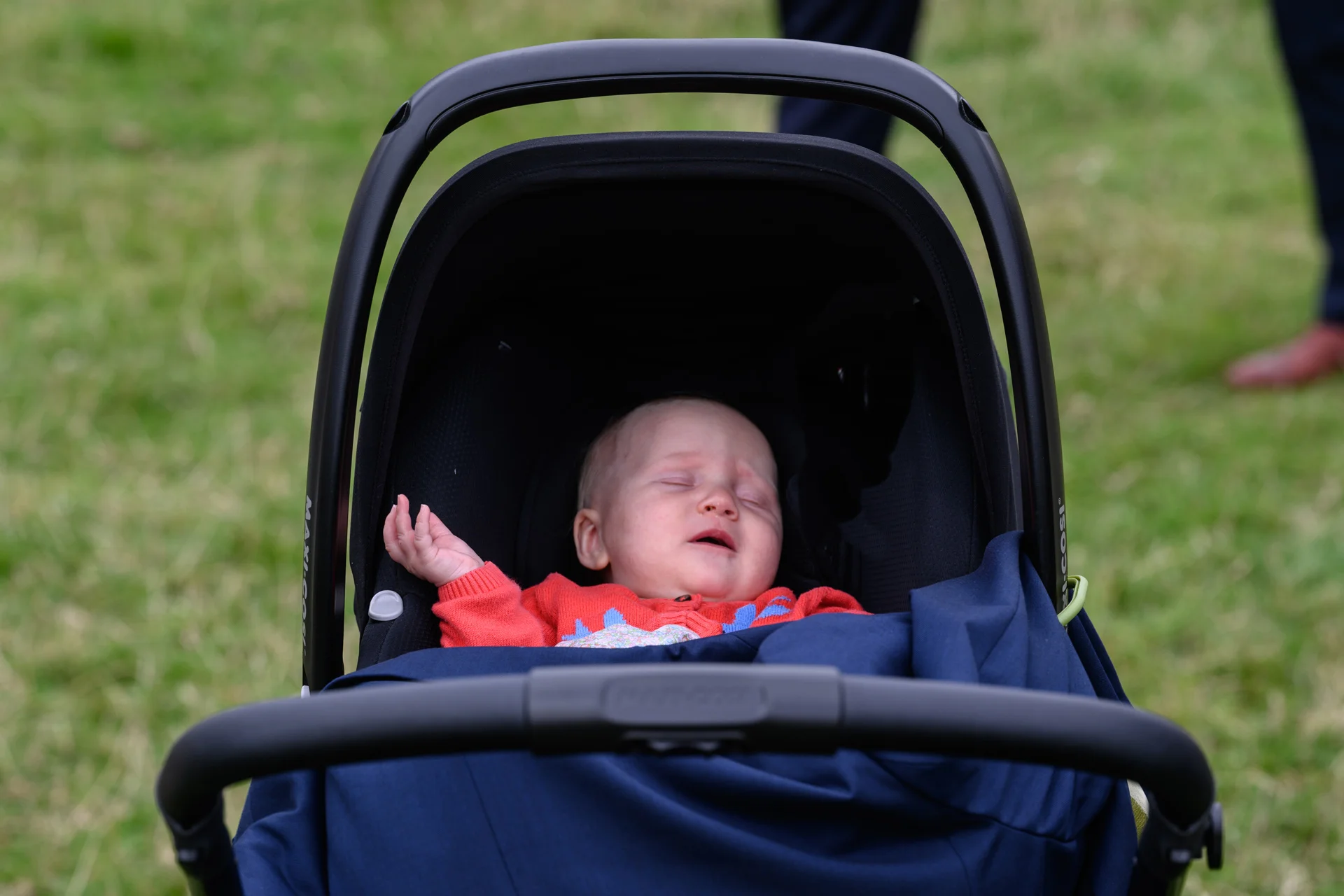 A tiny baby resting peacefully inside a black pushchair outdoors on a grassy lawn.
