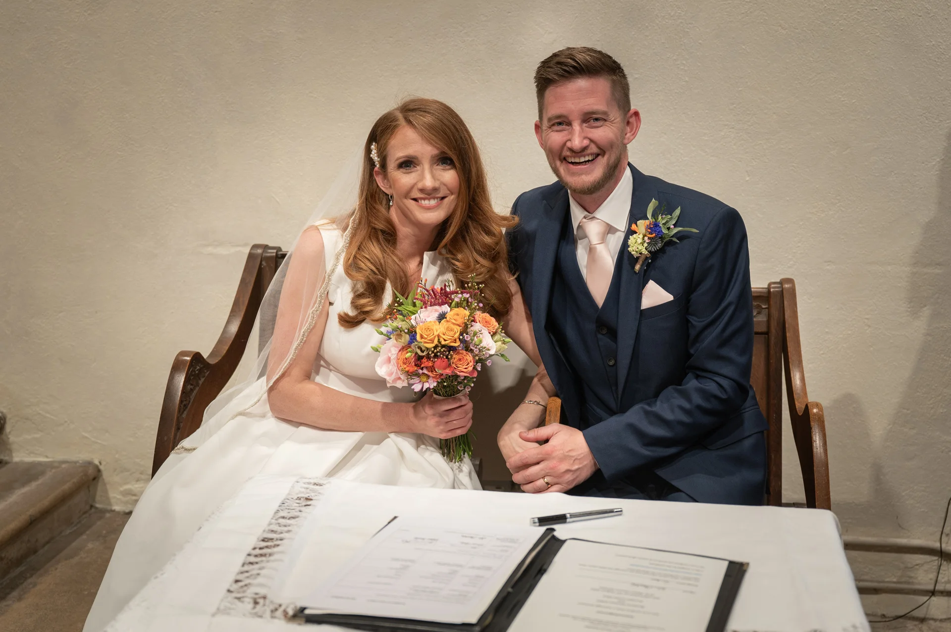 A smiling bride and groom sitting together at a table during their wedding ceremony, with the bride holding a colourful bouquet.