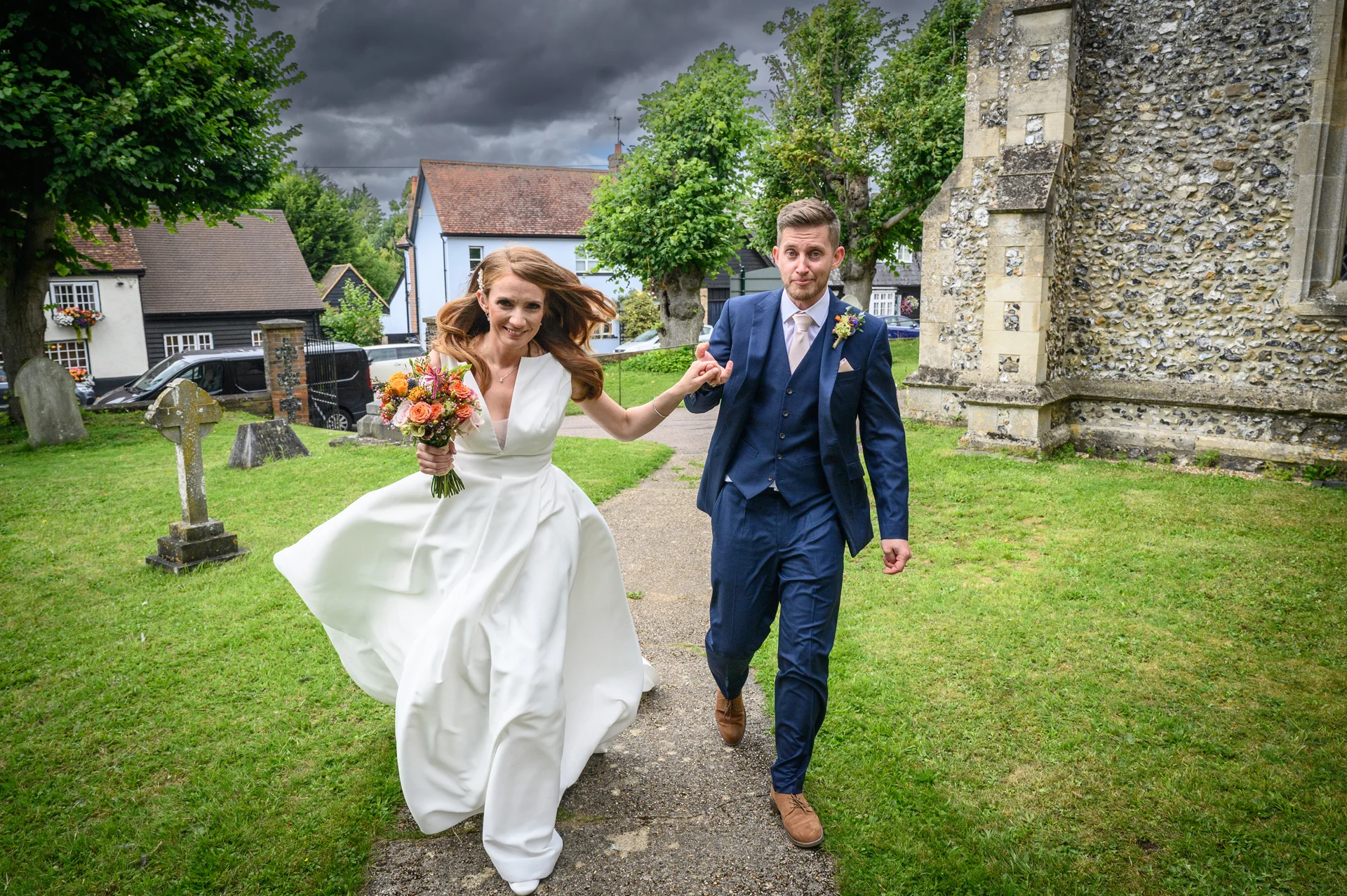 A newly married couple joyfully run and hold hands along a pathway outside a historic stone church under a dramatic, cloudy sky.