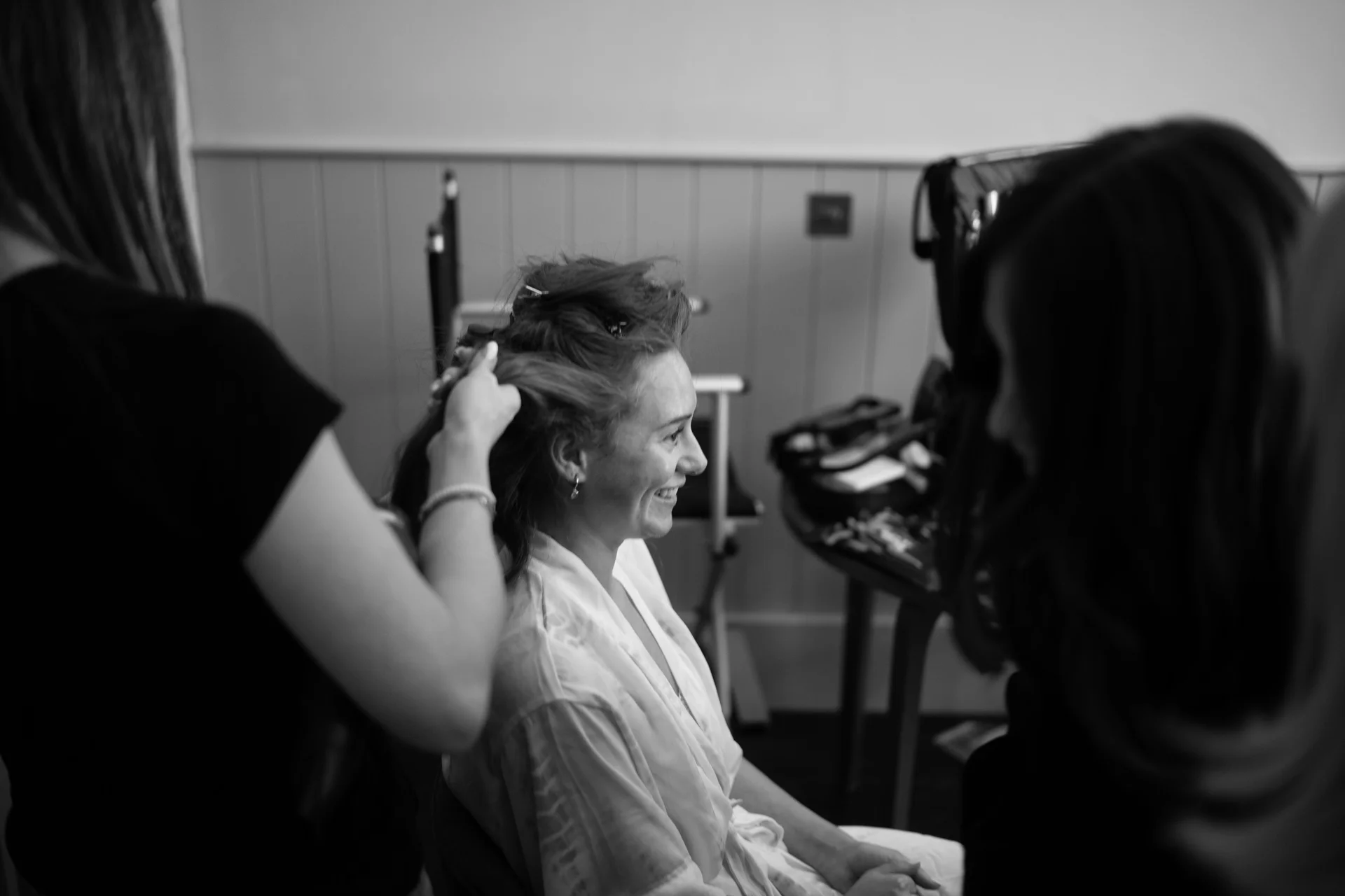 A smiling bride wearing a white robe has her hair styled during morning preparations while surrounded by members of the bridal party.