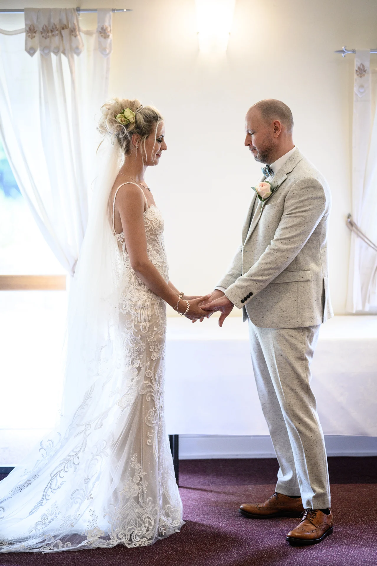 A bride in a lace fitted wedding dress and veil holds hands with her groom in a light grey suit during their indoor civil ceremony.