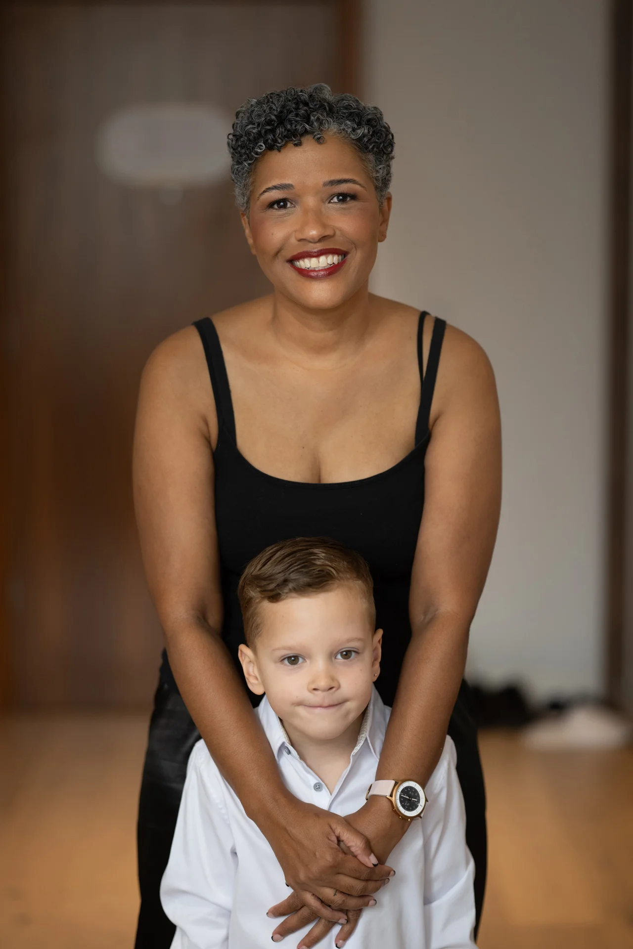 A smiling woman with short grey hair embraces a young boy wearing a white collared shirt indoors.