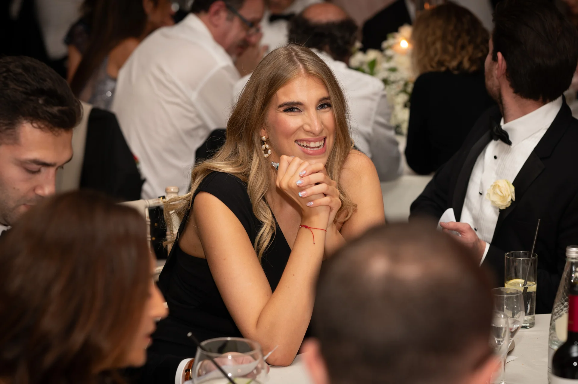 A smiling guest with long blonde hair rests her clasped hands on the dinner table during a lively wedding reception.
