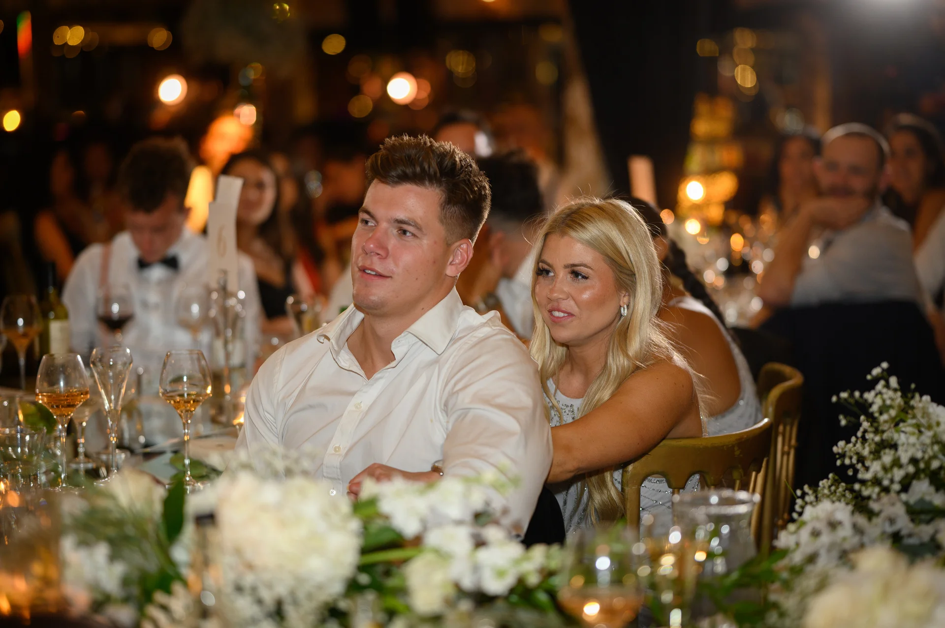 A couple seated at a candlelit wedding reception table laugh together during speeches, surrounded by white floral arrangements and glassware.
