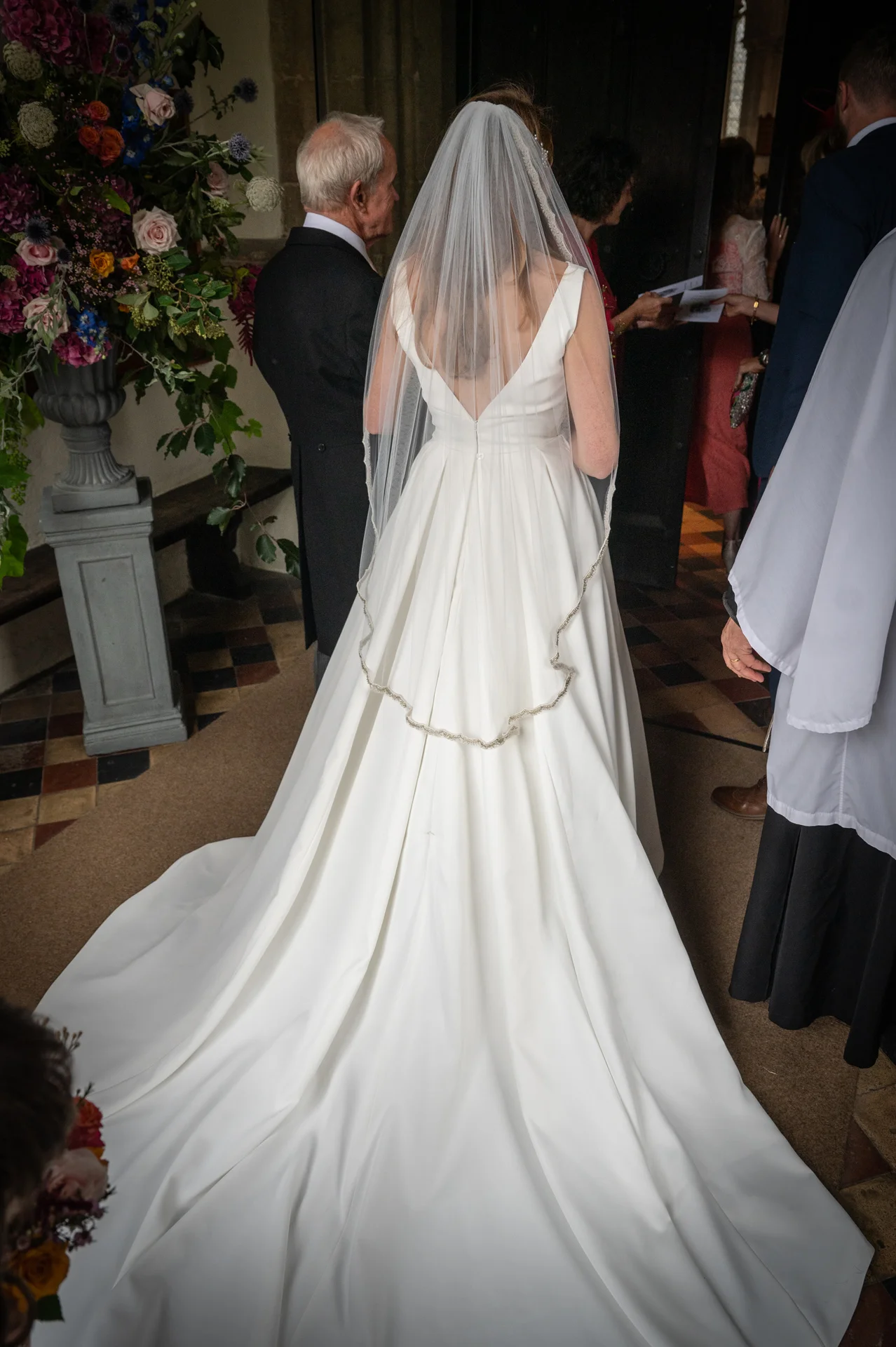 A bride wearing a satin A-line gown with a long train and lace-edged veil stands at the church entrance with her father.