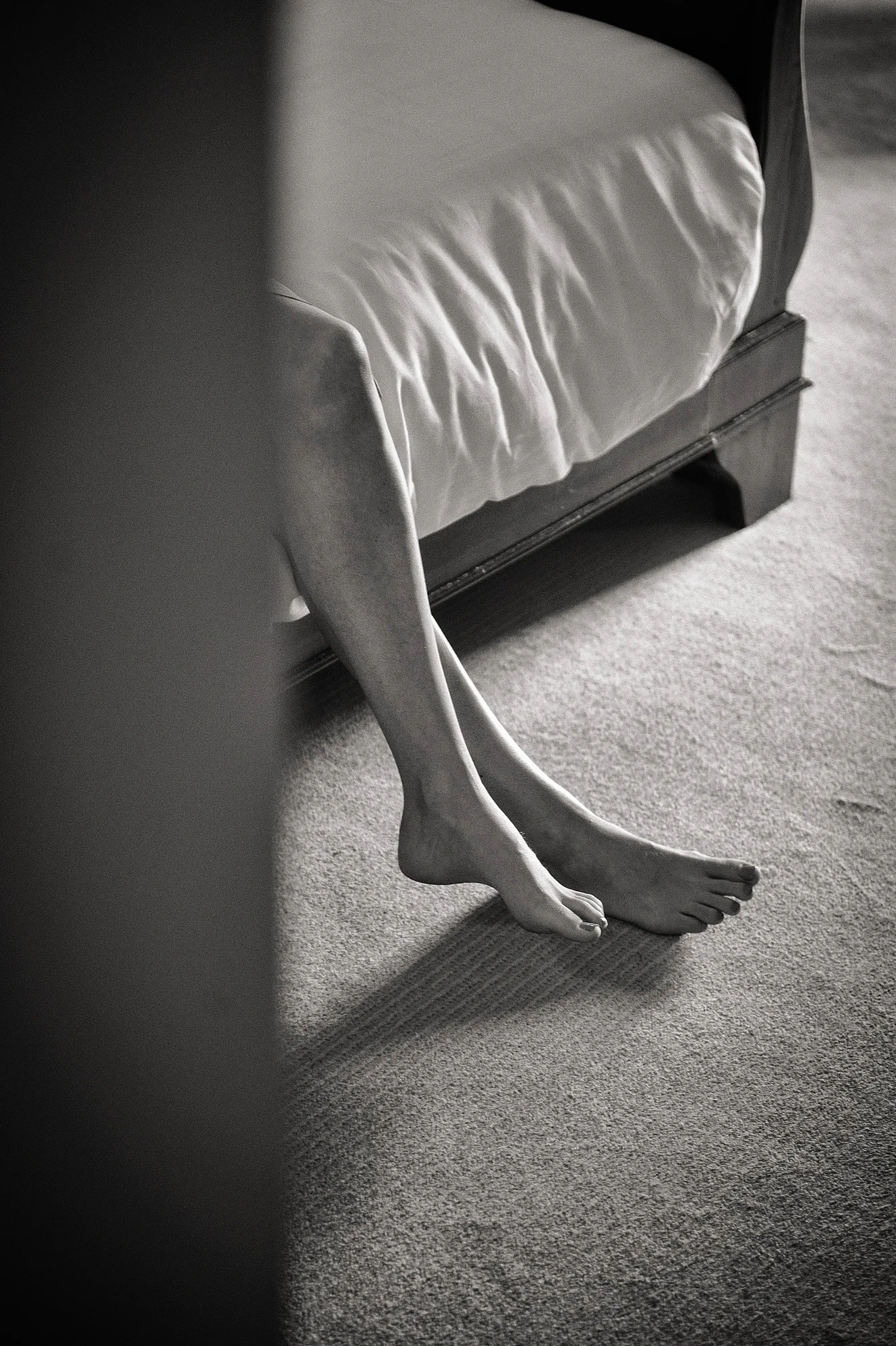 A candid black and white shot captures a barefoot bride relaxing by the edge of a bed during morning preparations.