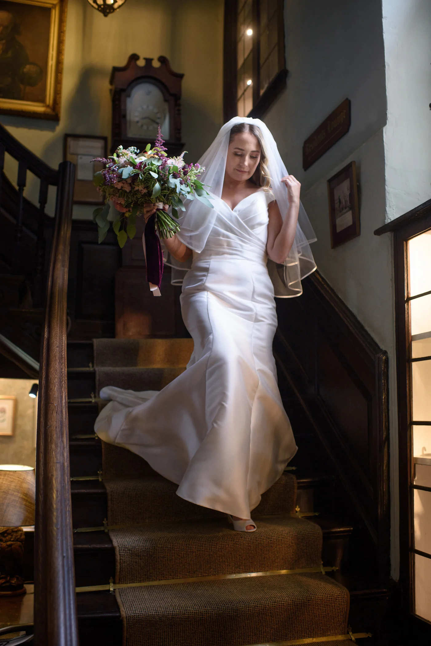 A graceful bride descends a grand wooden staircase inside a traditional venue, holding an oversized cascading bouquet and wearing an elegant satin v-neck gown with a chapel veil.