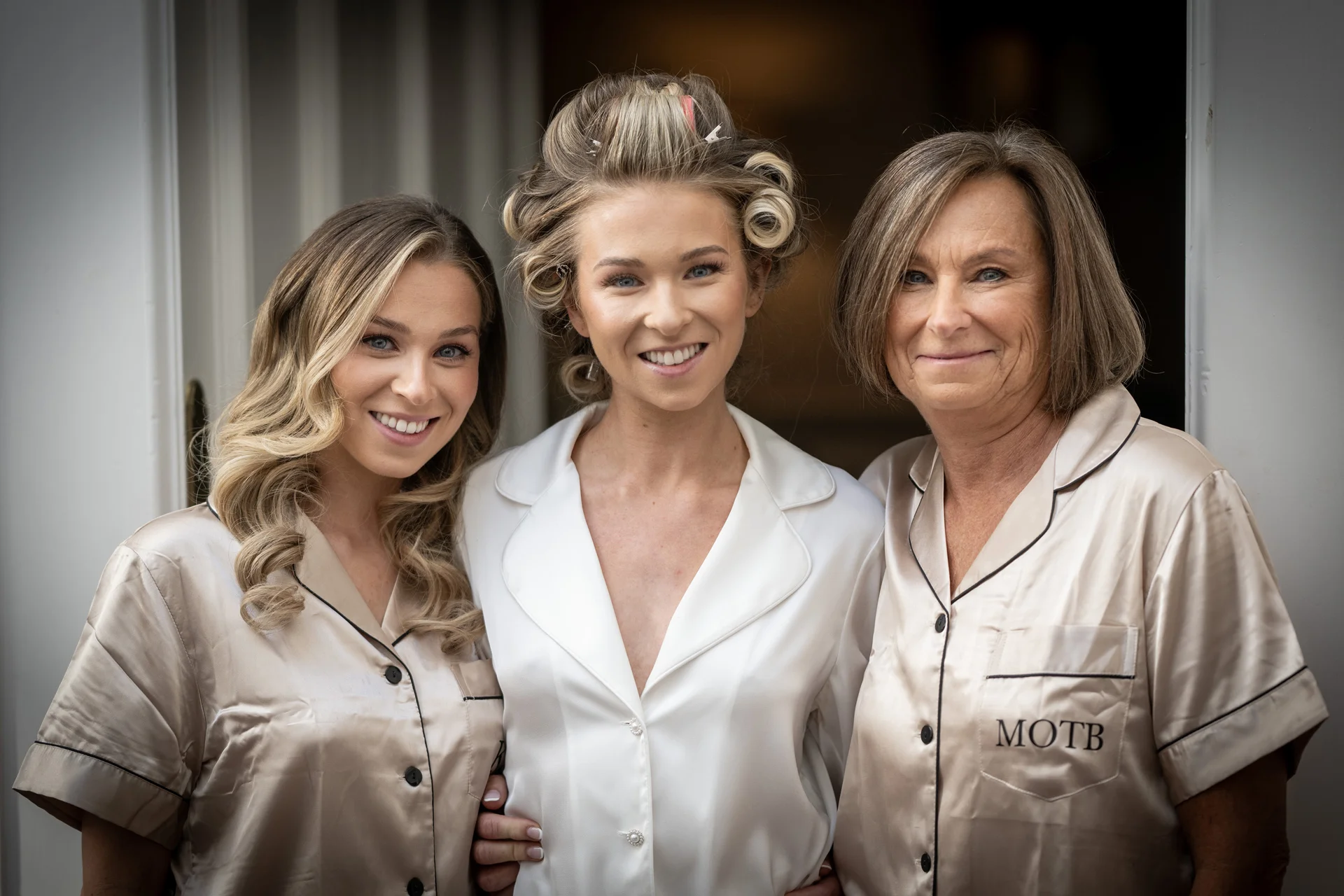 A bride with hair rollers in her hair stands smiling between a bridesmaid and her mother, all wearing coordinating getting ready pyjamas.