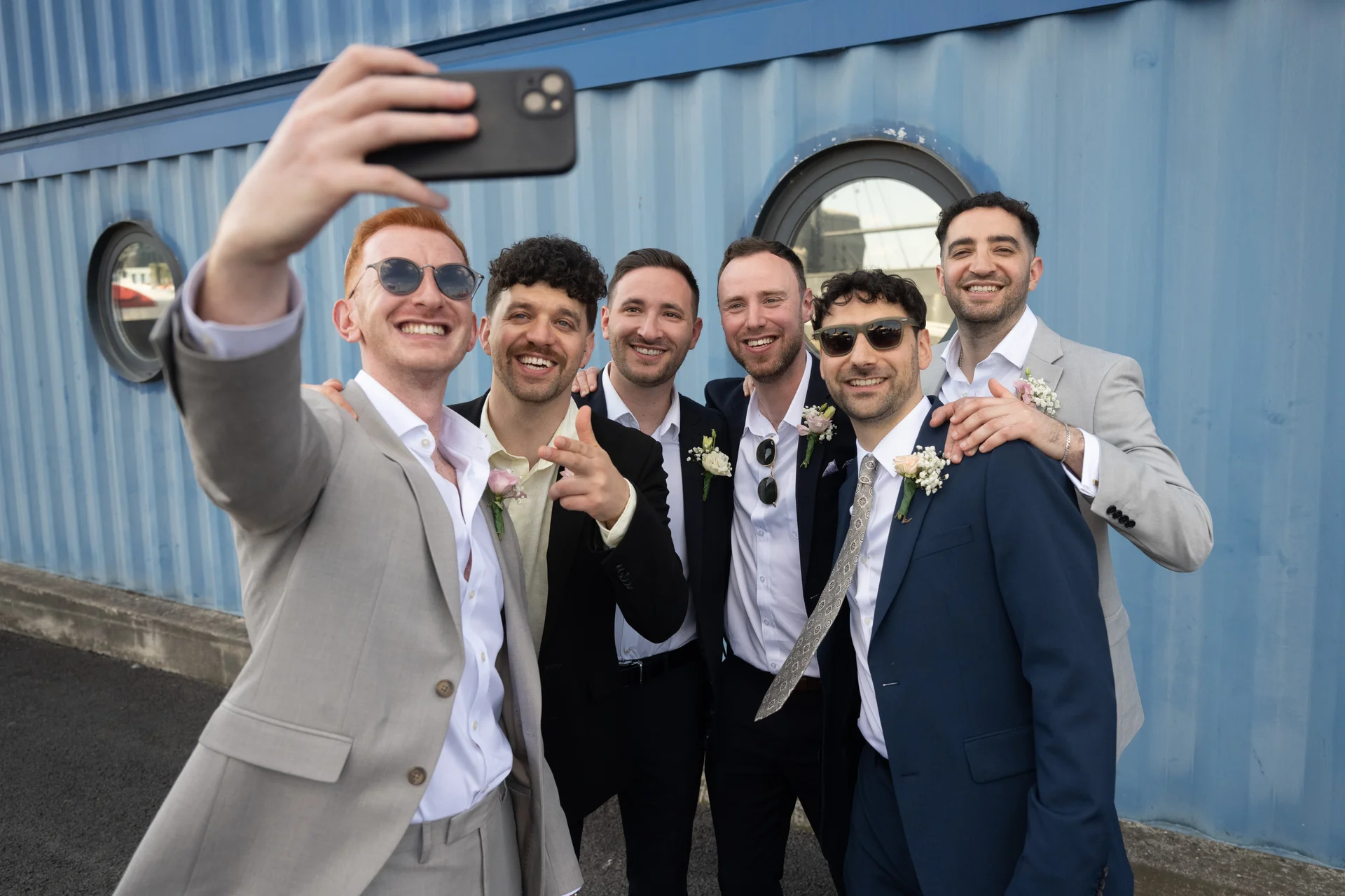A group of smiling groomsmen in smart suits take a selfie together in front of a blue shipping container wall.