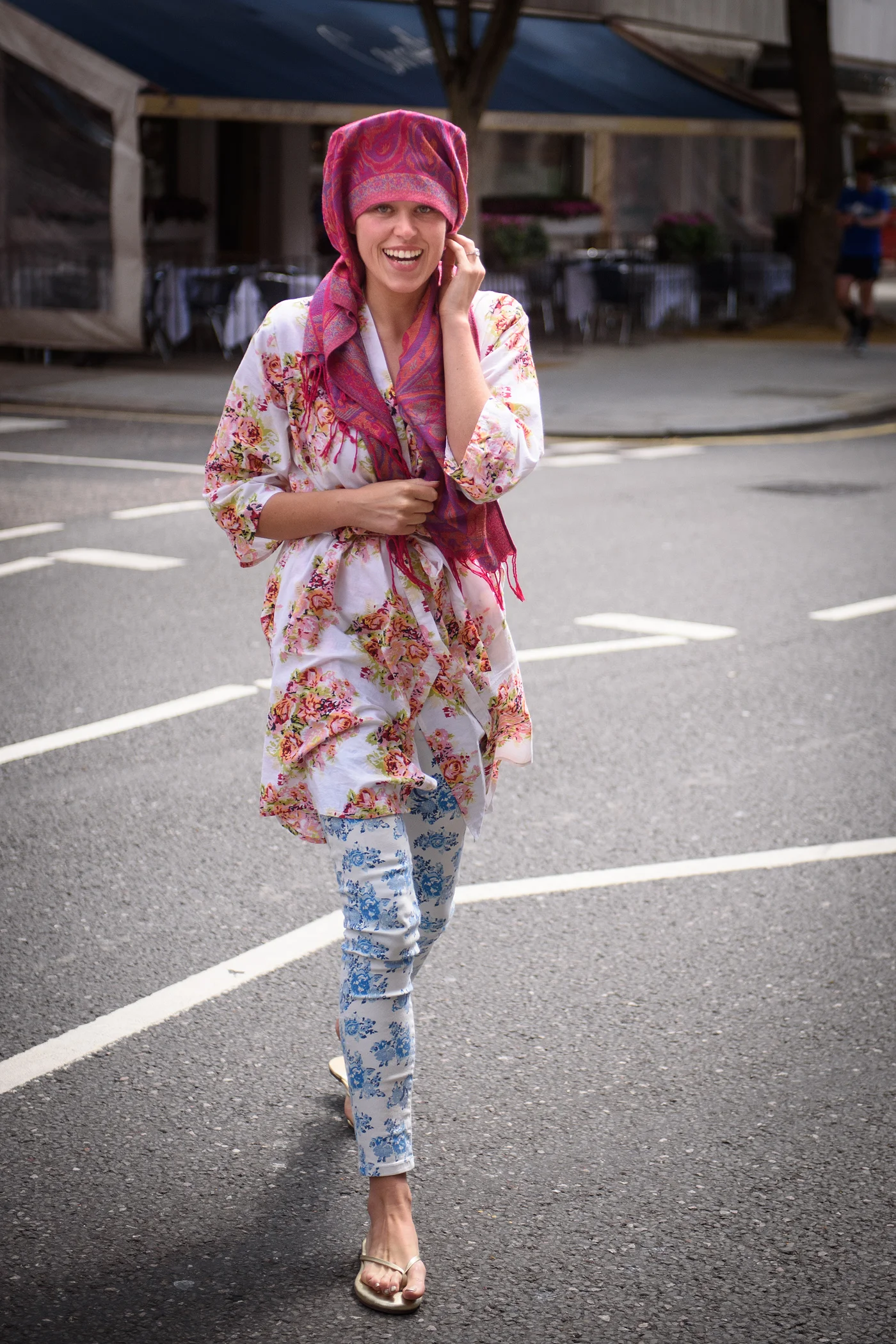 A joyful bride wearing a floral robe and a pink headscarf walks smiling along a city street during bridal preparations.