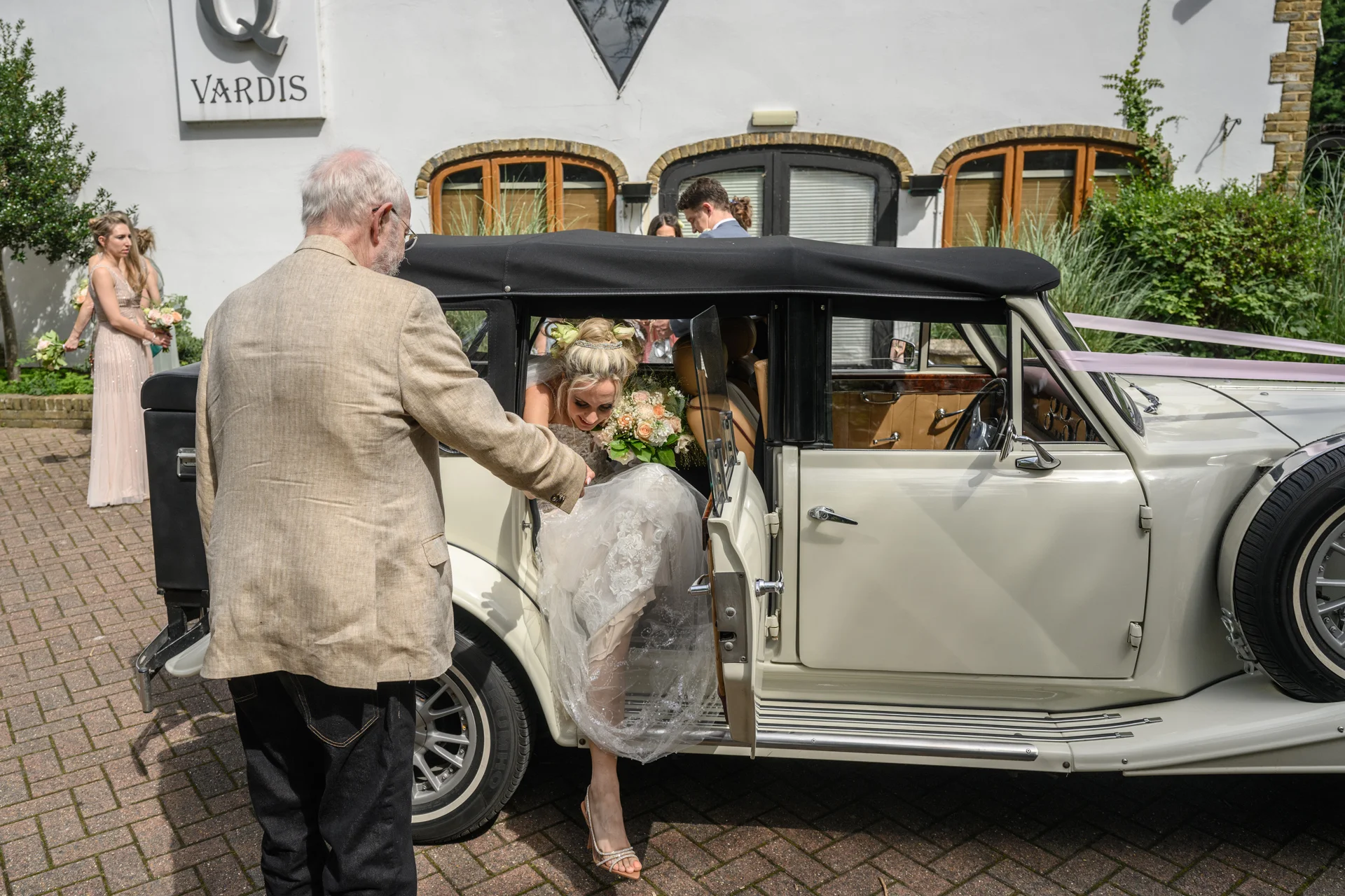 A smiling bride steps out of a classic vintage wedding car, assisted by her father outside a white building with a cobbled courtyard.