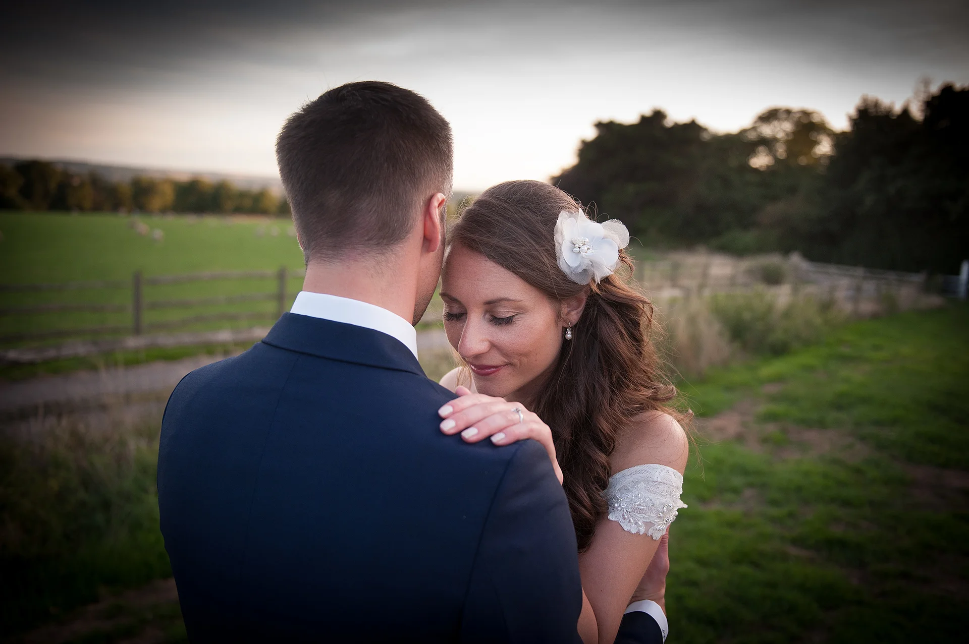 A newlywed couple embraces tenderly outdoors in the countryside at dusk, with the bride resting her hand on the groom's shoulder.