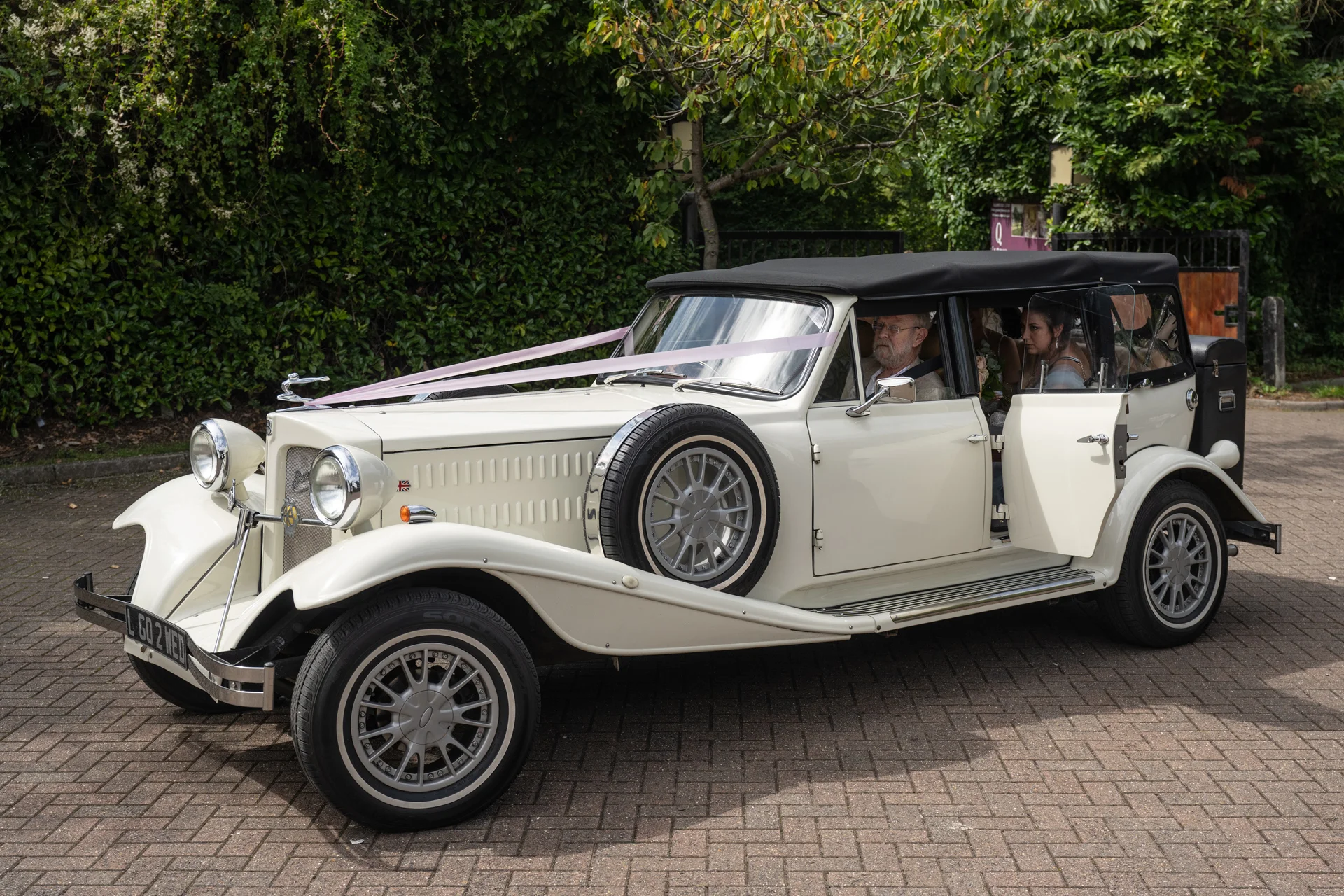 A vintage cream wedding car decorated with pink ribbons is parked on a paved driveway with passengers visible inside.