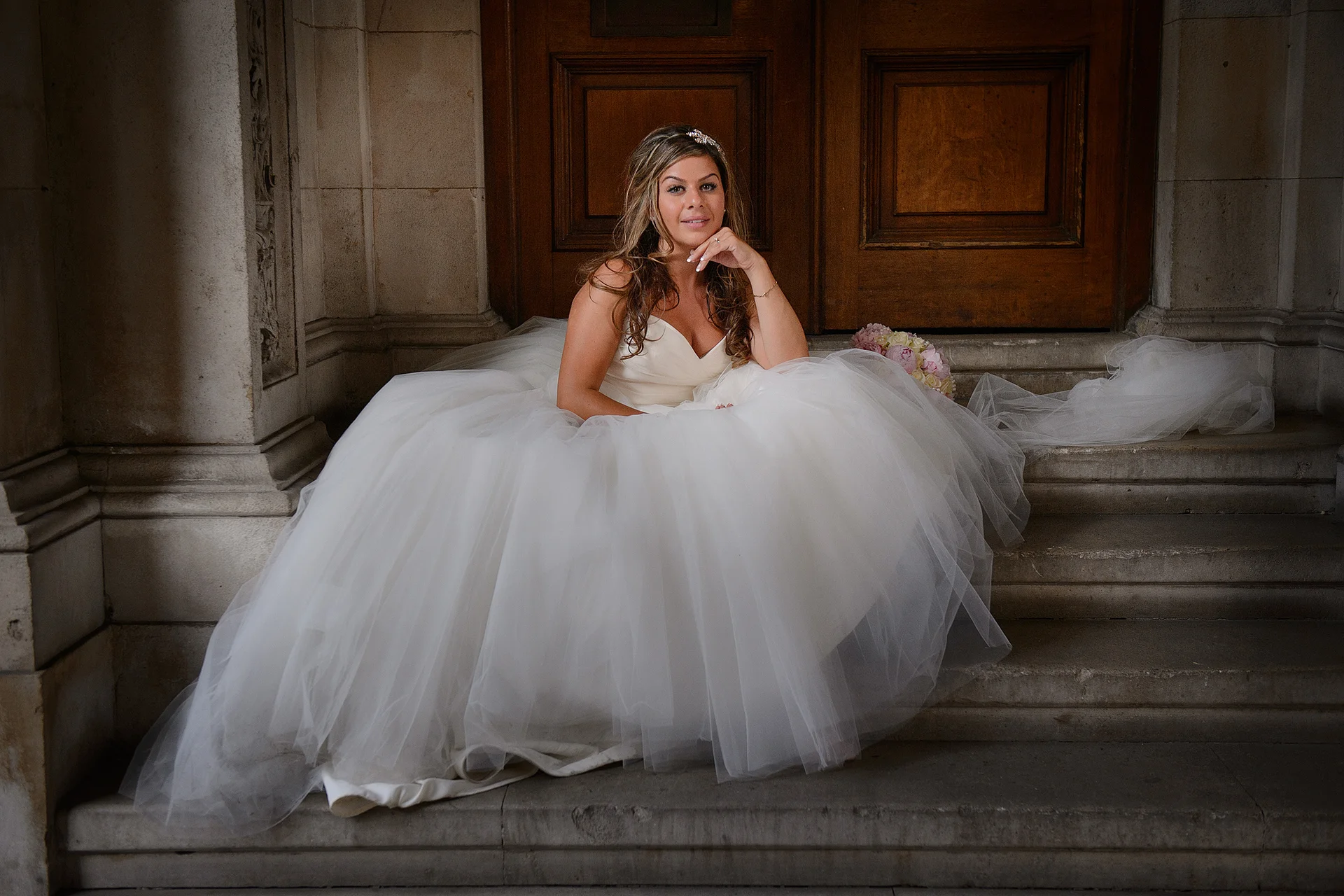 A smiling bride wearing a voluminous tulle ball gown sits gracefully on stone steps in front of heavy wooden doors, holding a bouquet.