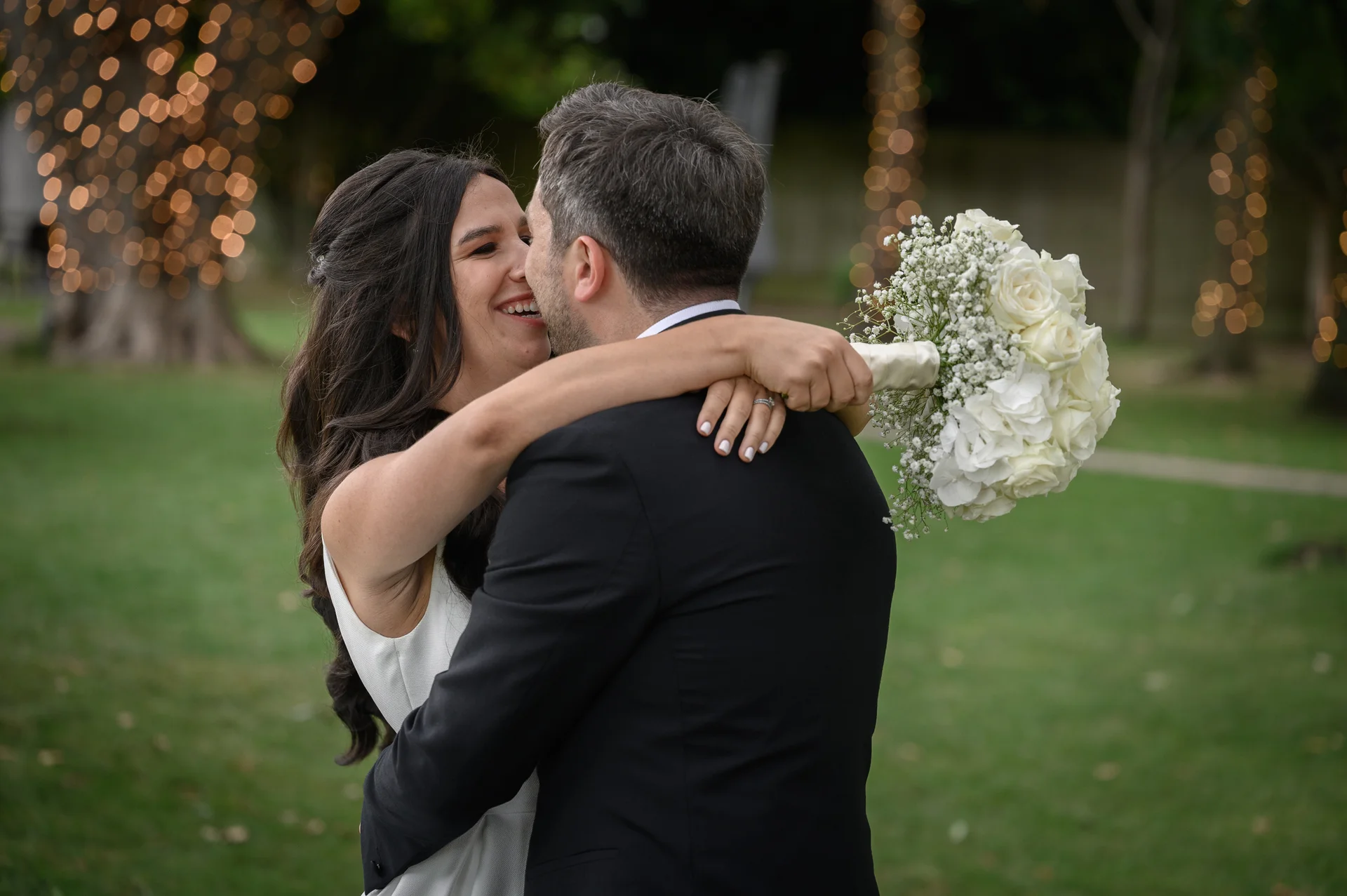 A joyful bride and groom embrace outdoors against a backdrop of trees wrapped in twinkling fairy lights, with the bride holding a white rose and baby's breath bouquet.