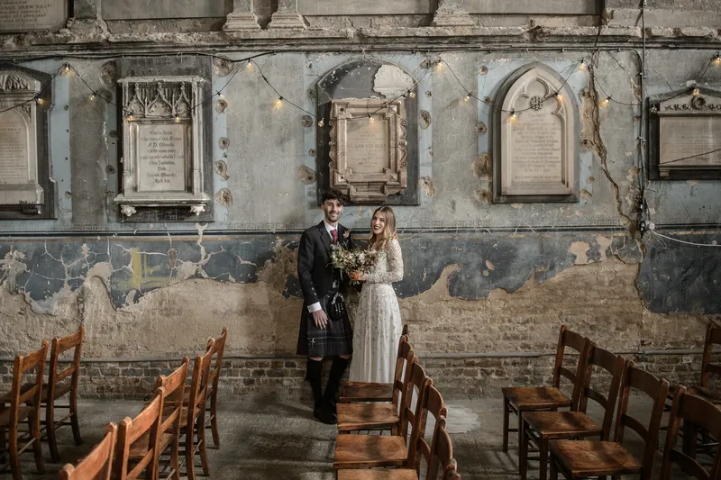 A newlywed couple poses together inside a historic chapel with weathered stone walls, distressed plaster, and rows of vintage wooden chairs.