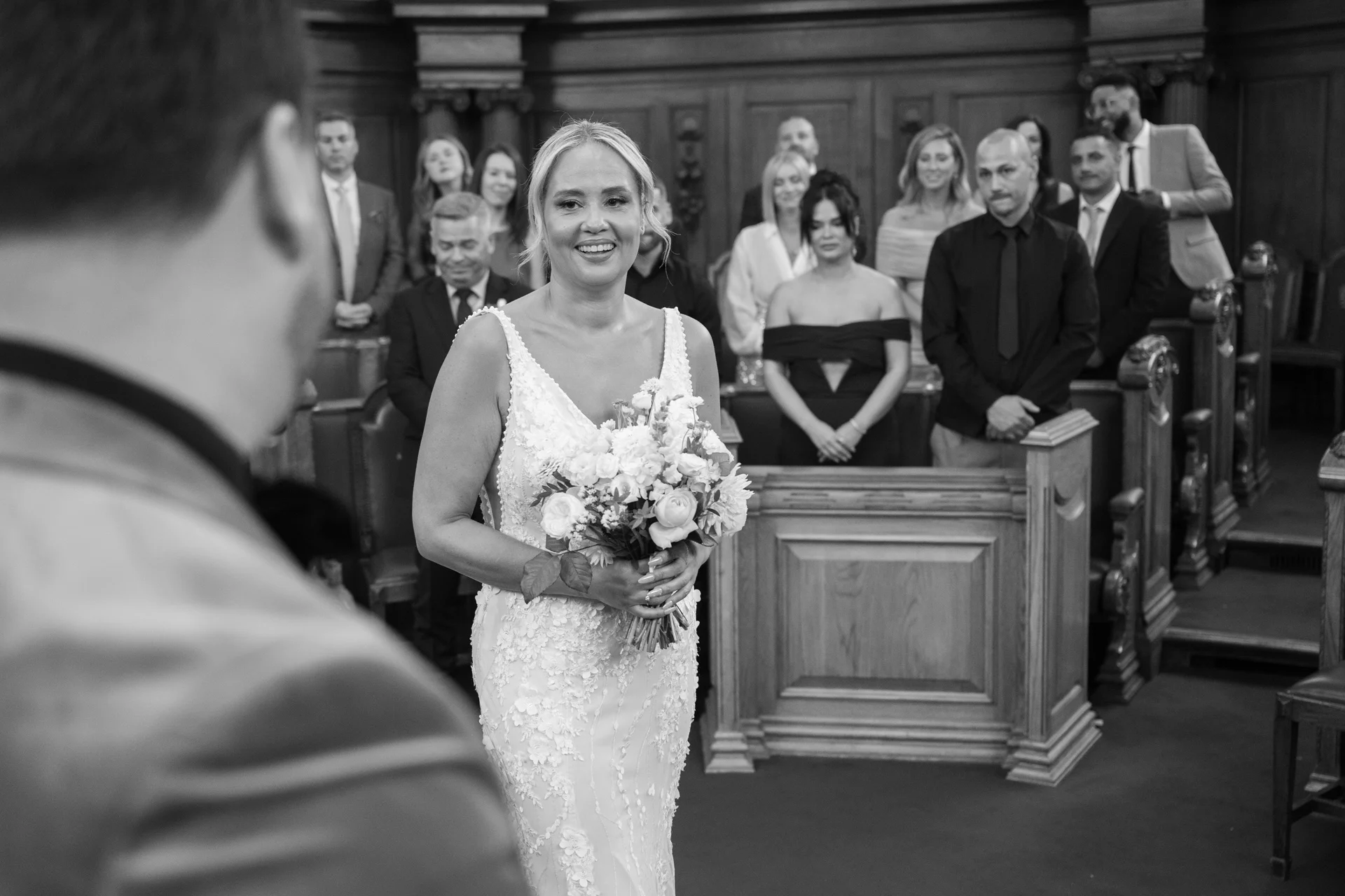 A smiling bride holding a floral bouquet walks down the aisle towards the groom during an indoor wedding ceremony.