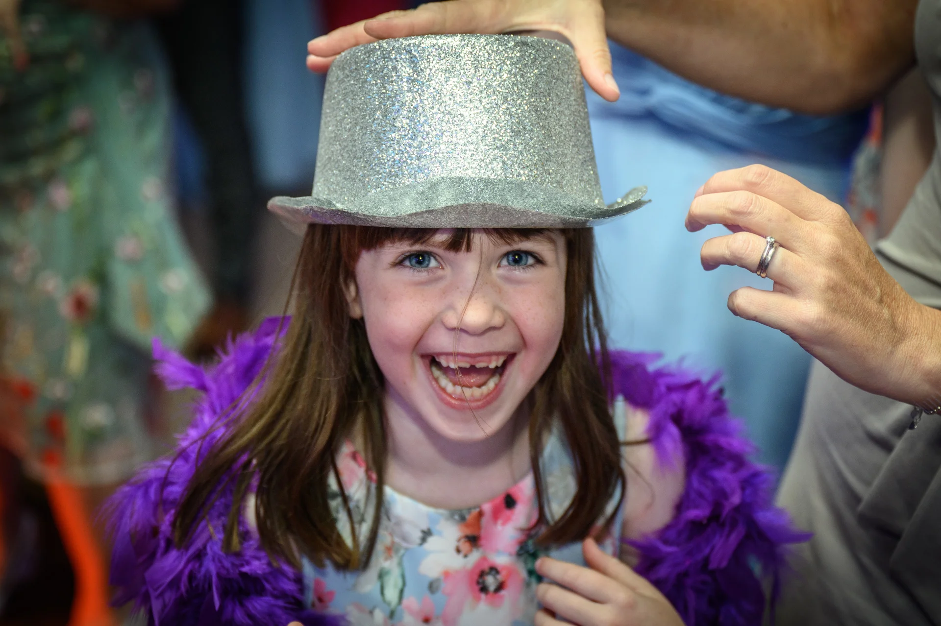 A young flower girl laughs joyfully while wearing a sparkly silver top hat and a vibrant purple feather boa at a wedding reception.