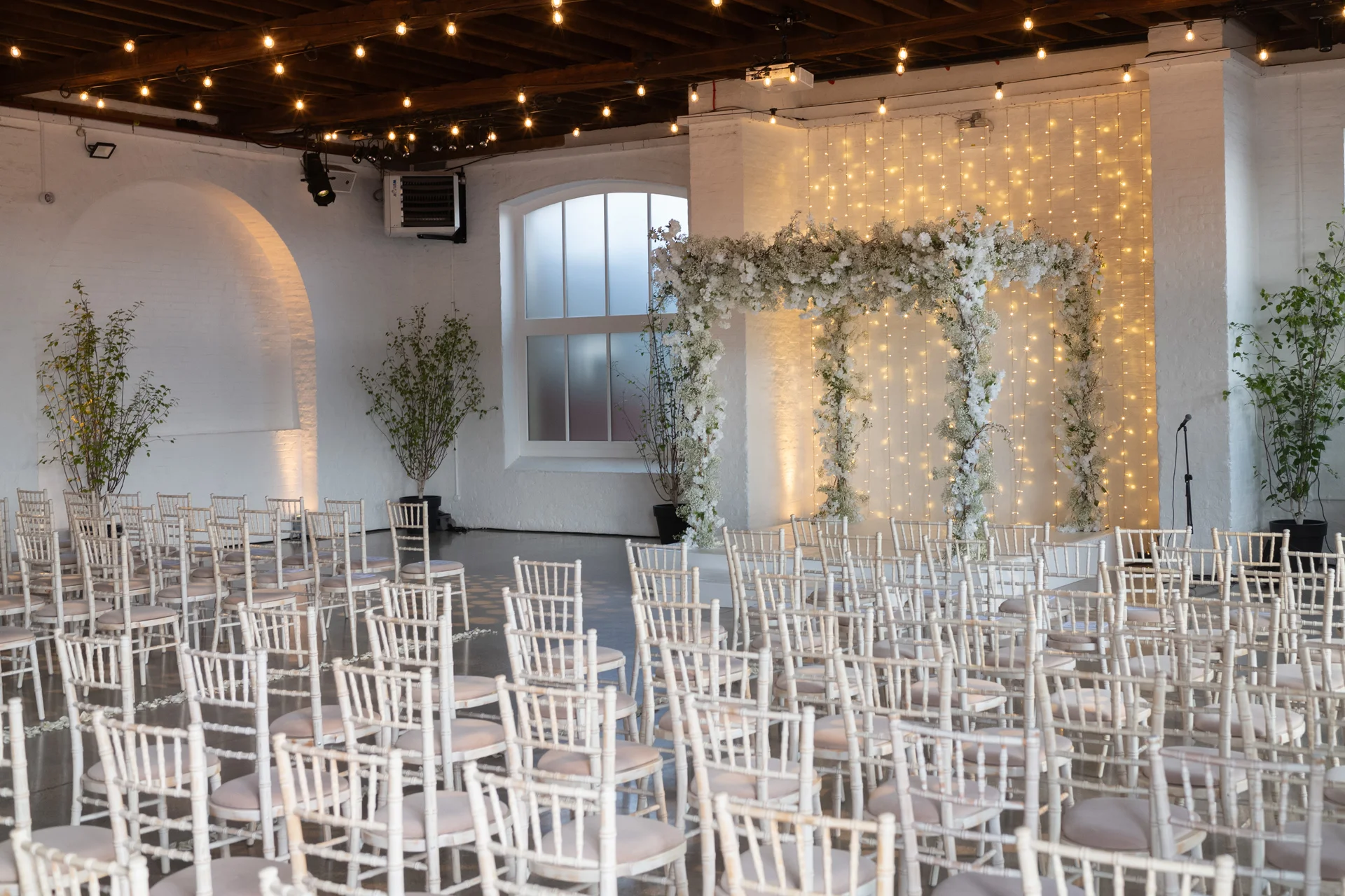 A brightly lit industrial wedding venue featuring rows of white chiavari chairs facing a stunning floral ceremony arch backed by a warm fairy light wall.