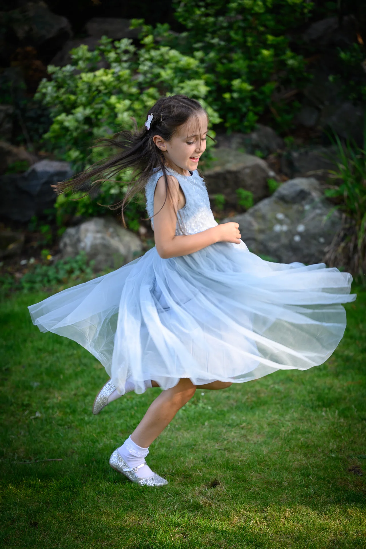 A young flower girl in a pale blue tulle dress twirls happily on a green lawn with her hair caught in motion.