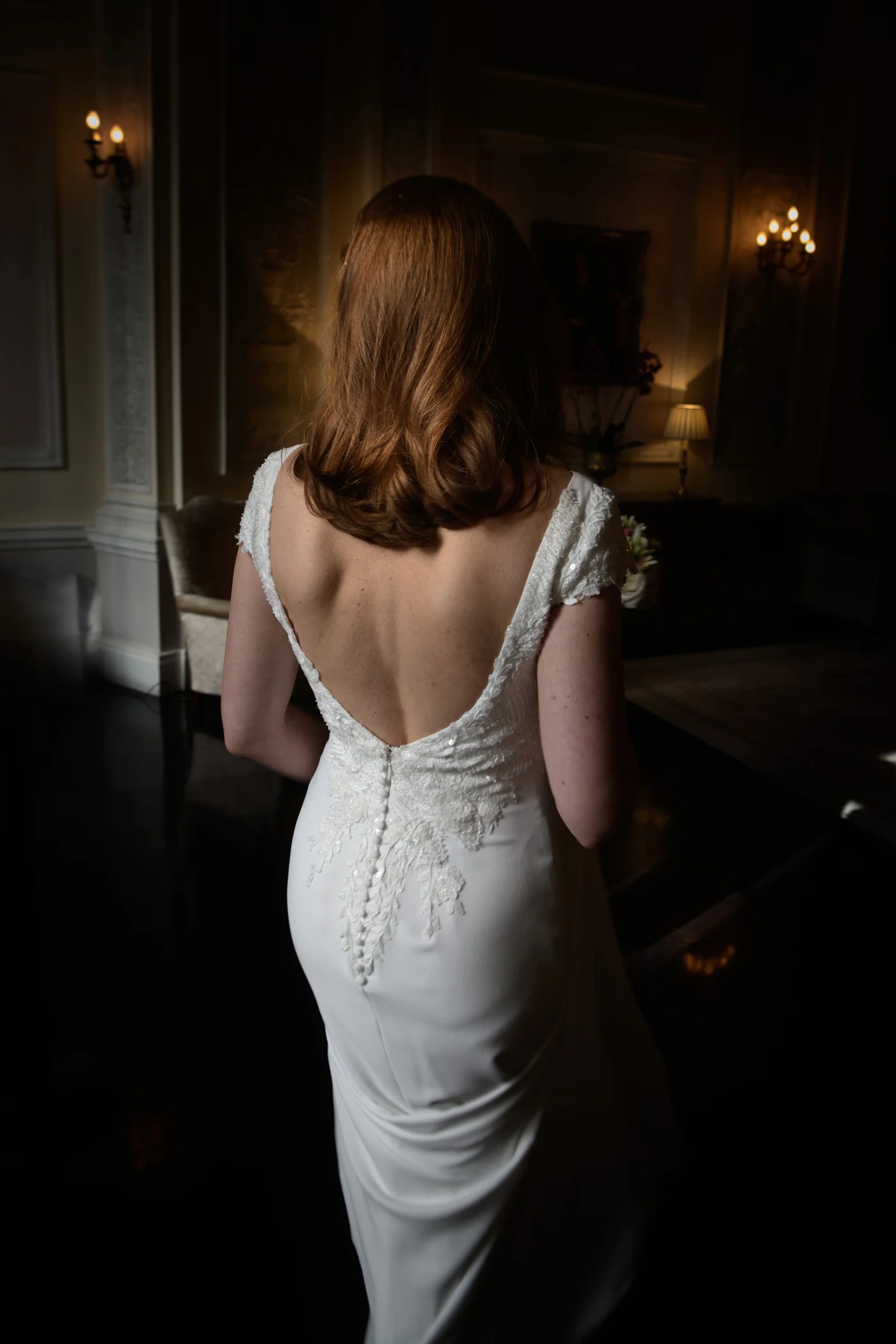 A bride stands facing away, showcasing the low back and delicate button detailing of her elegant wedding gown inside a dimly lit historic room.