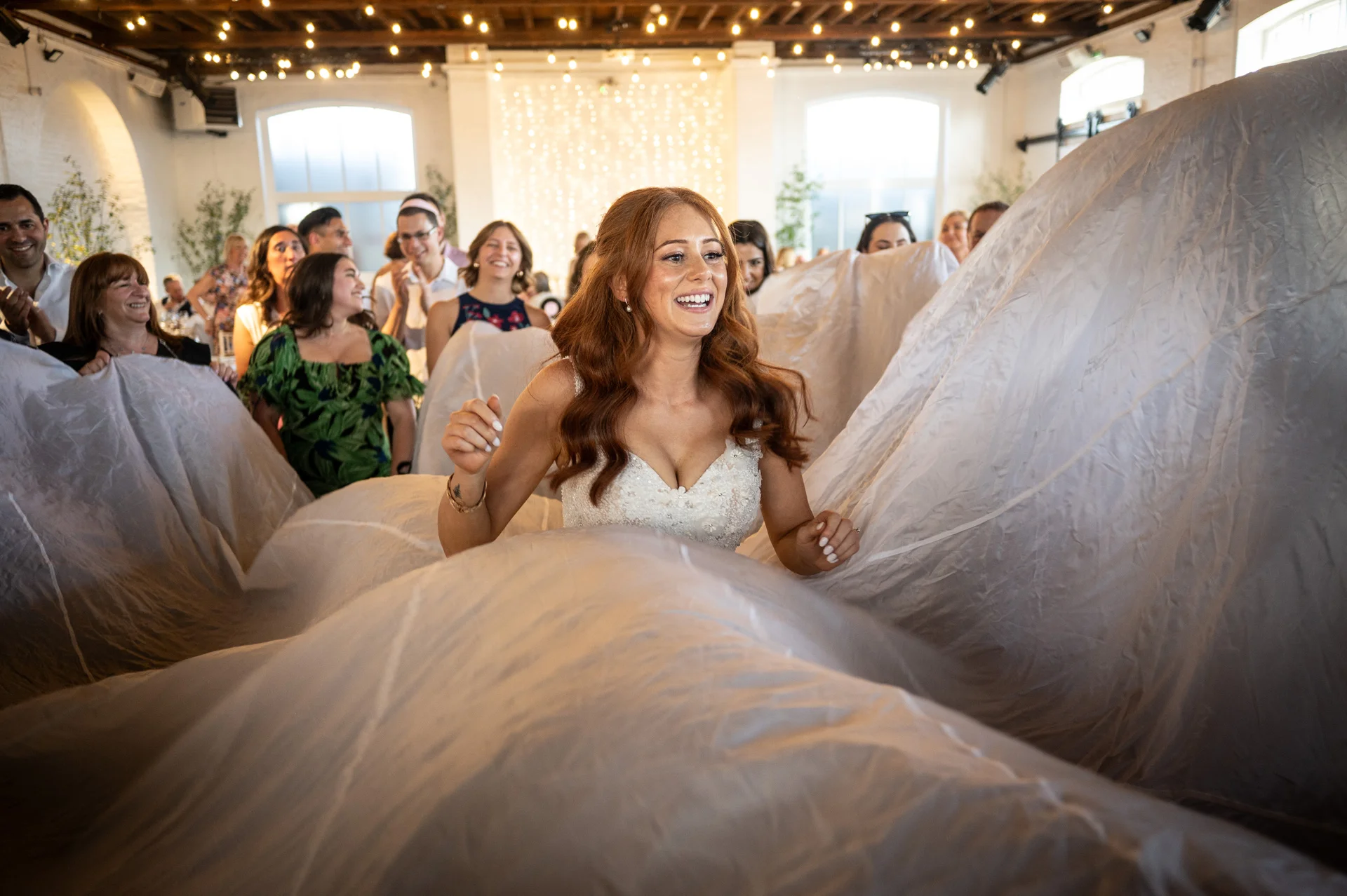 A joyful red-haired bride laughs while emerging from underneath a large white parachute cloth inside a rustic barn wedding venue lit by fairy lights.