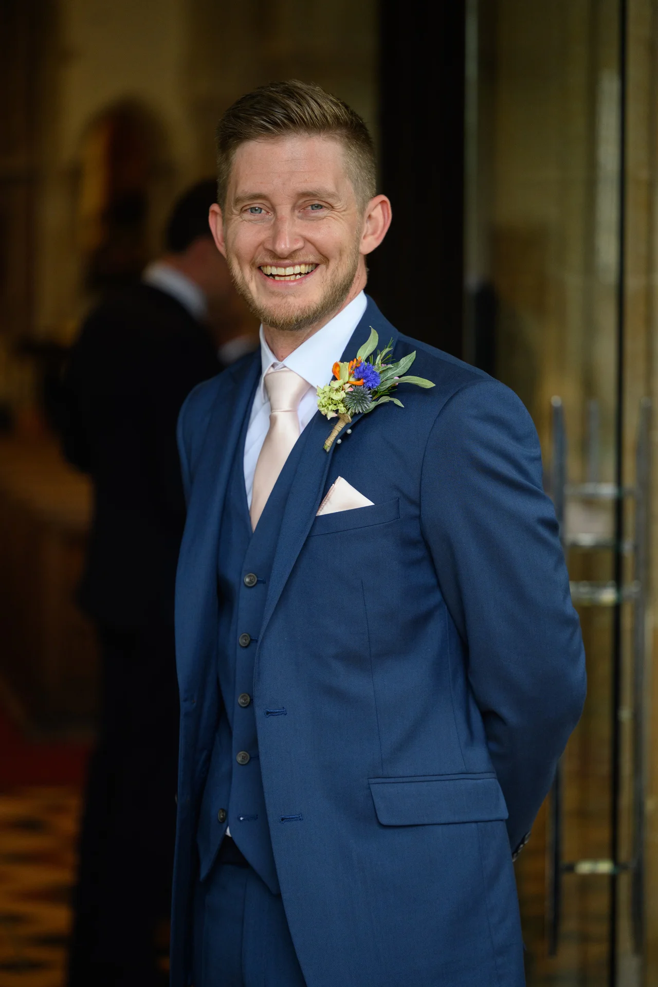 A smiling groom wearing a smart navy three-piece suit, pink tie and floral buttonhole stands posing for a portrait.