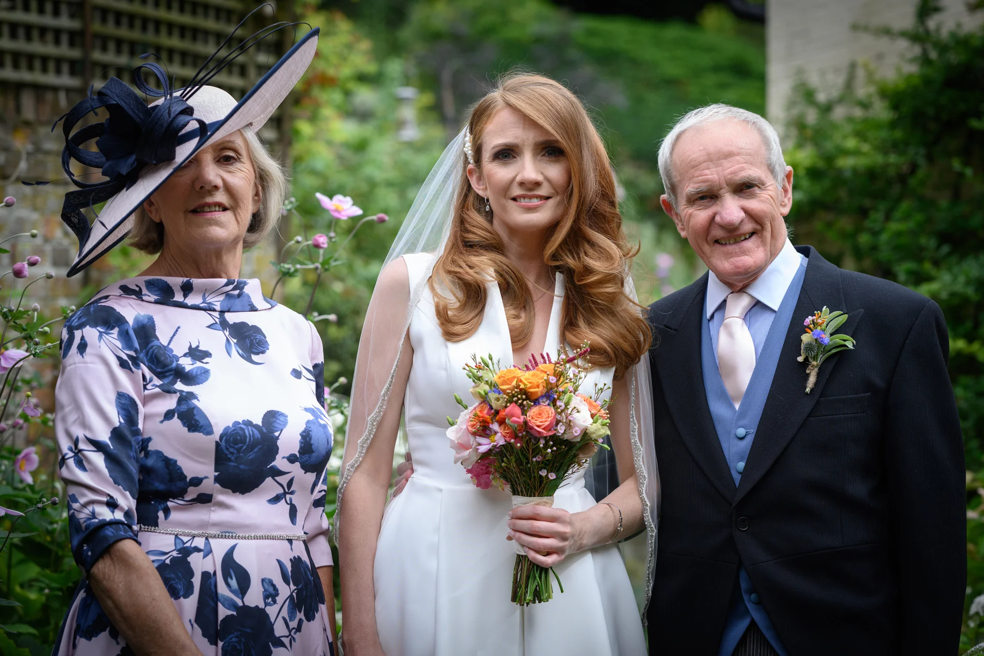 A smiling bride holding a colourful bouquet stands outdoors in a garden flanked by her mother in a floral dress and father in a morning suit.