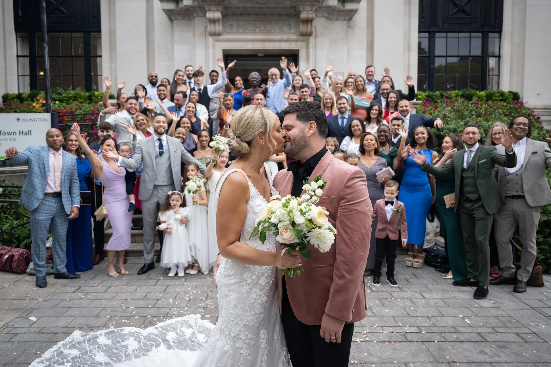 A newlywed couple share a kiss outside a historic town hall steps while a large crowd of wedding guests cheer and wave in the background.