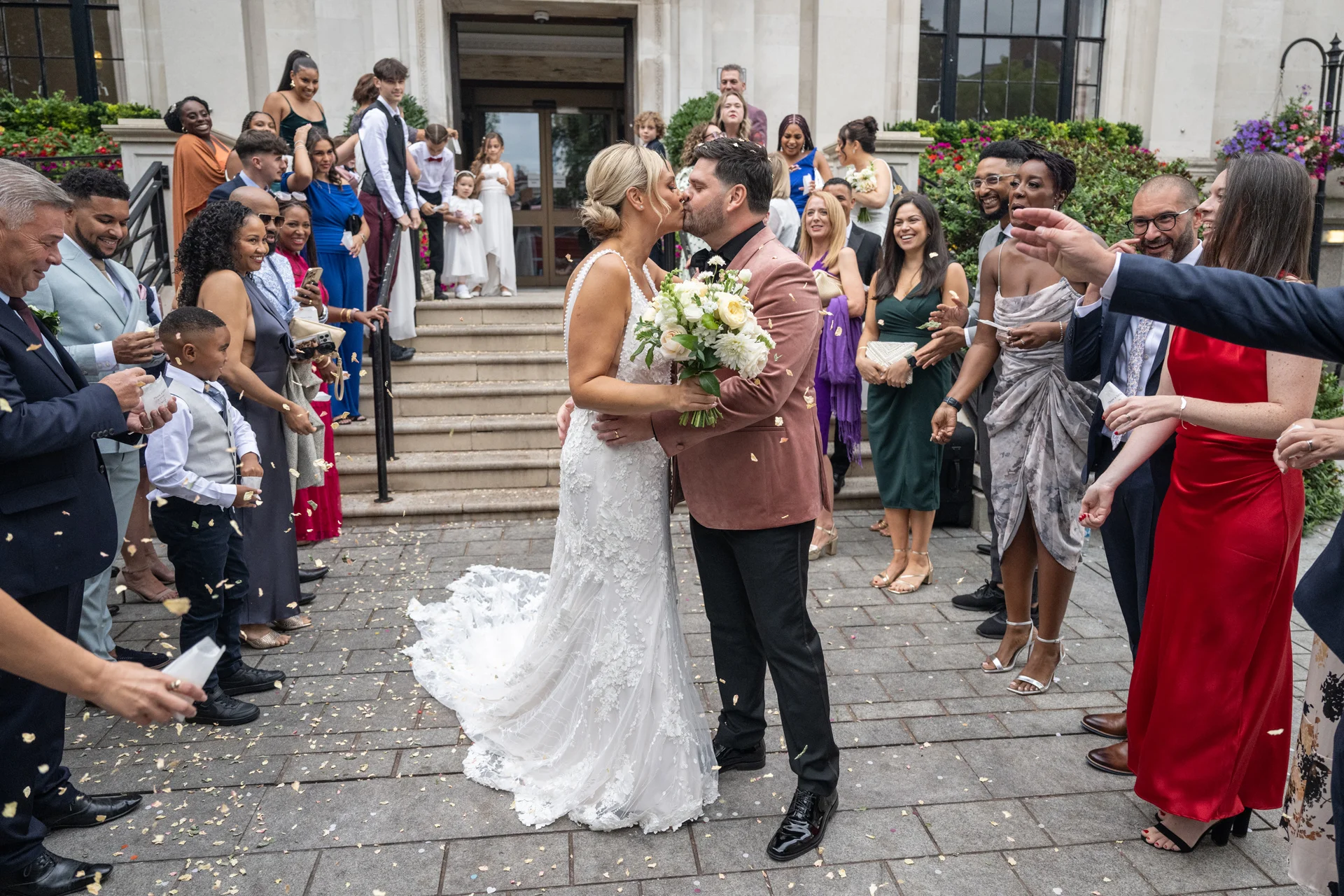 Newlyweds share their first kiss outside a venue while smiling wedding guests shower them with confetti.