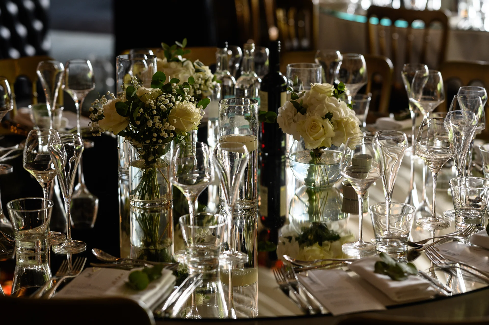 An elegantly styled wedding reception tablescape featuring a mirrored surface, white roses, delicate greenery, crystal glassware, and neatly folded napkins with custom menus.