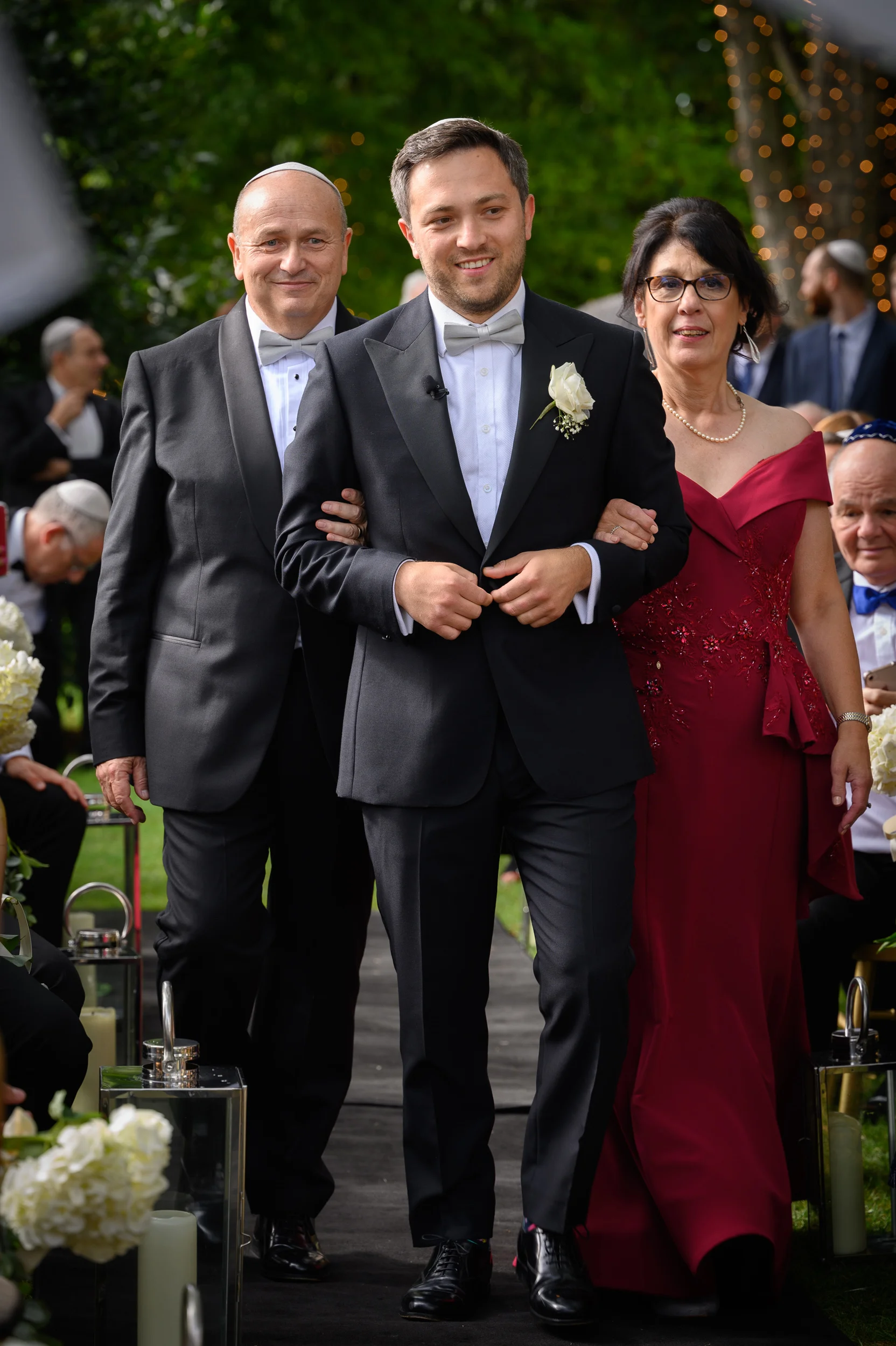 A smiling groom walks down the ceremony aisle linked arm-in-arm with his mother in a burgundy gown and his father in a classic tuxedo.