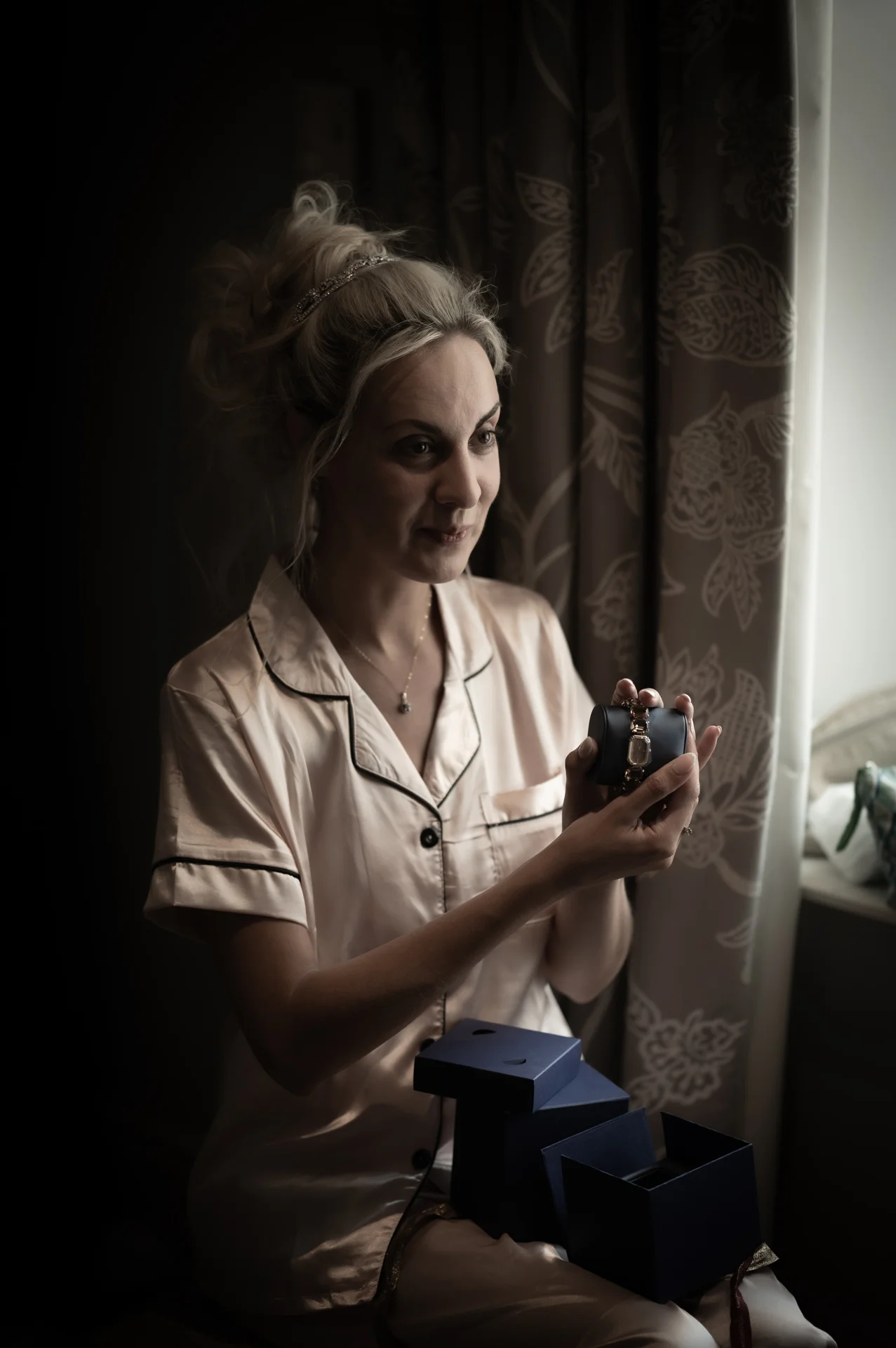 A blonde bride wearing matching silk pyjamas looks lovingly at a watch gift while sitting by a window during bridal preparations.