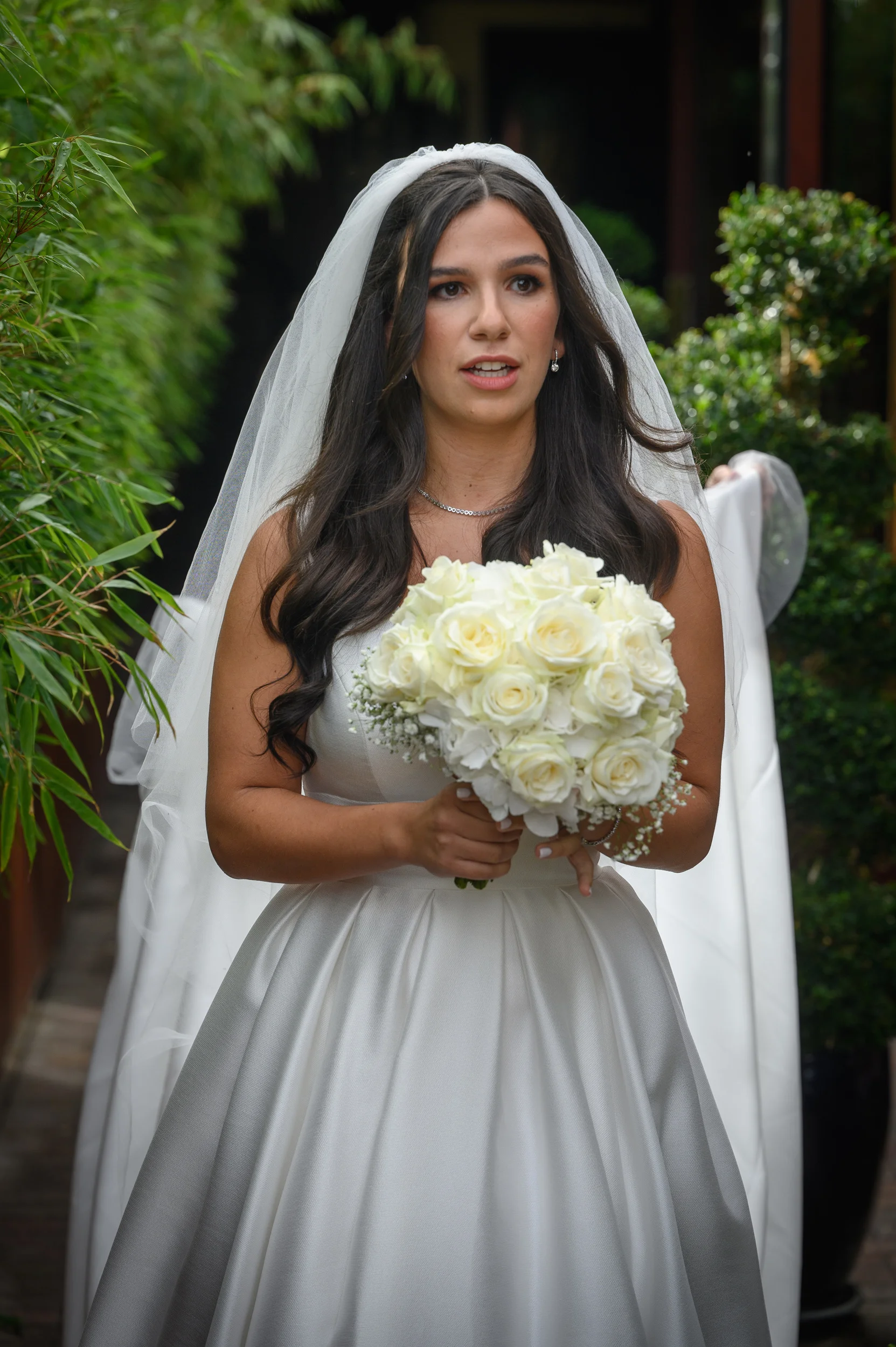 A stunning bride with long dark hair and a veil holds a lush white rose bouquet while wearing an A-line satin gown outdoors.