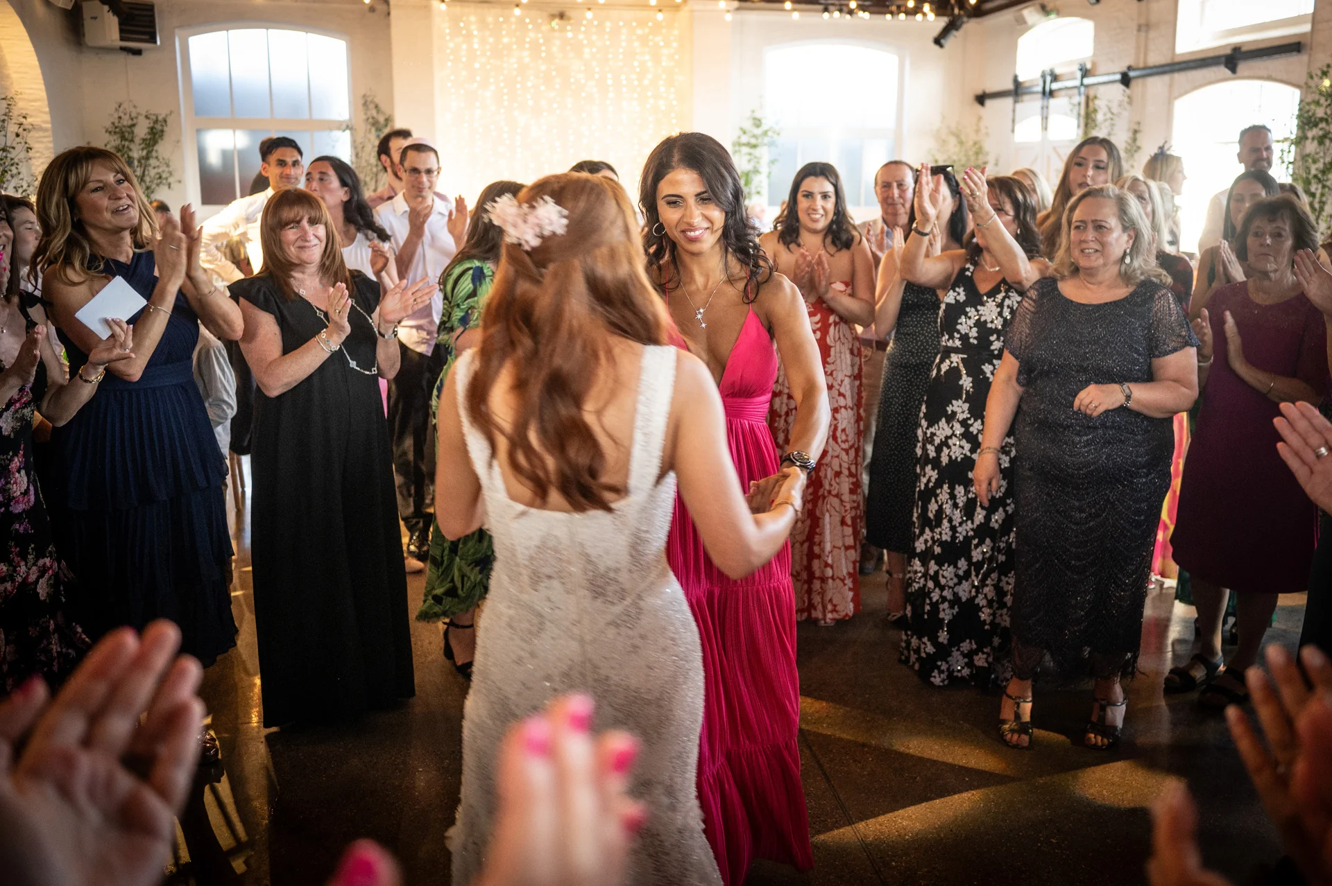Two brides share a joyful first dance surrounded by cheering wedding guests inside an industrial venue illuminated by a fairy light canopy.